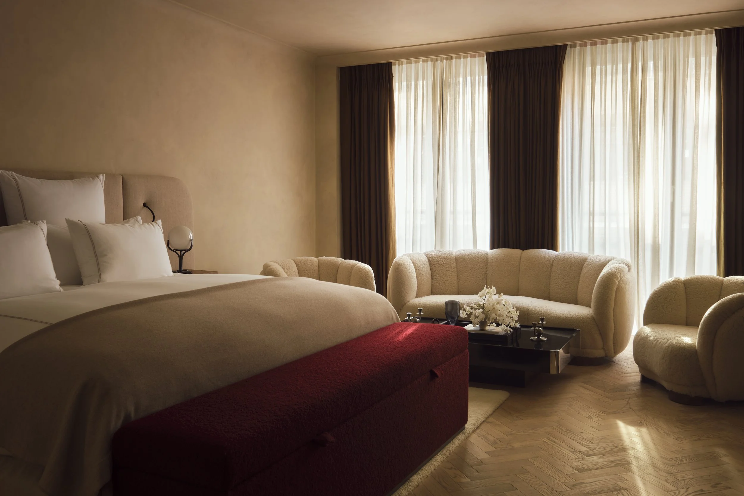 A hotel room featuring a bed with white pillows and beige bedding, a sitting area with beige curved couches, a black coffee table with floral arrangements. St Clement London