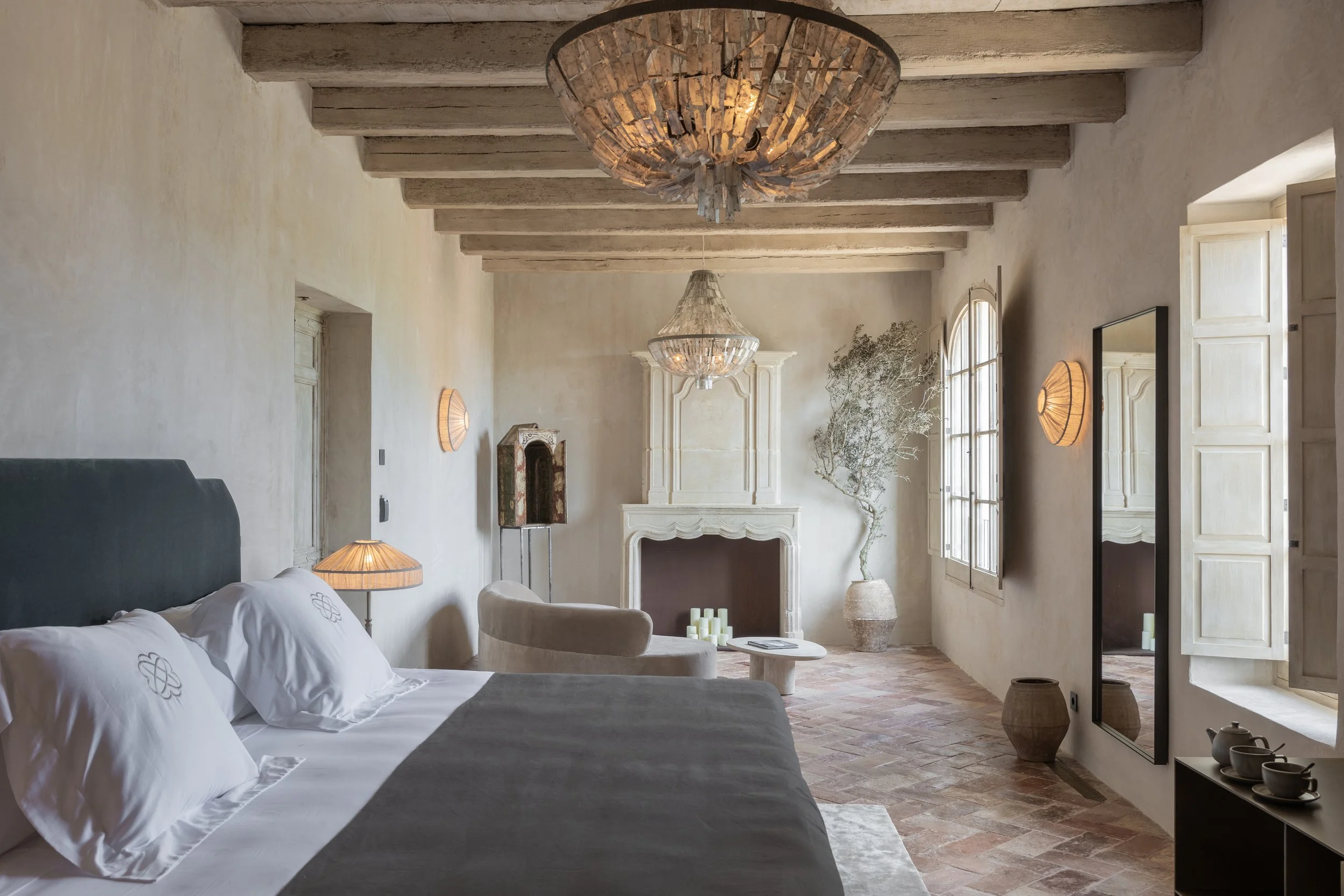 Elegant bedroom with exposed wooden beams ceiling, fireplace, large window, decorative plants, mirrors, and minimalist furniture in neutral tones. Vestige Collection Son Vell Menorca Spain
