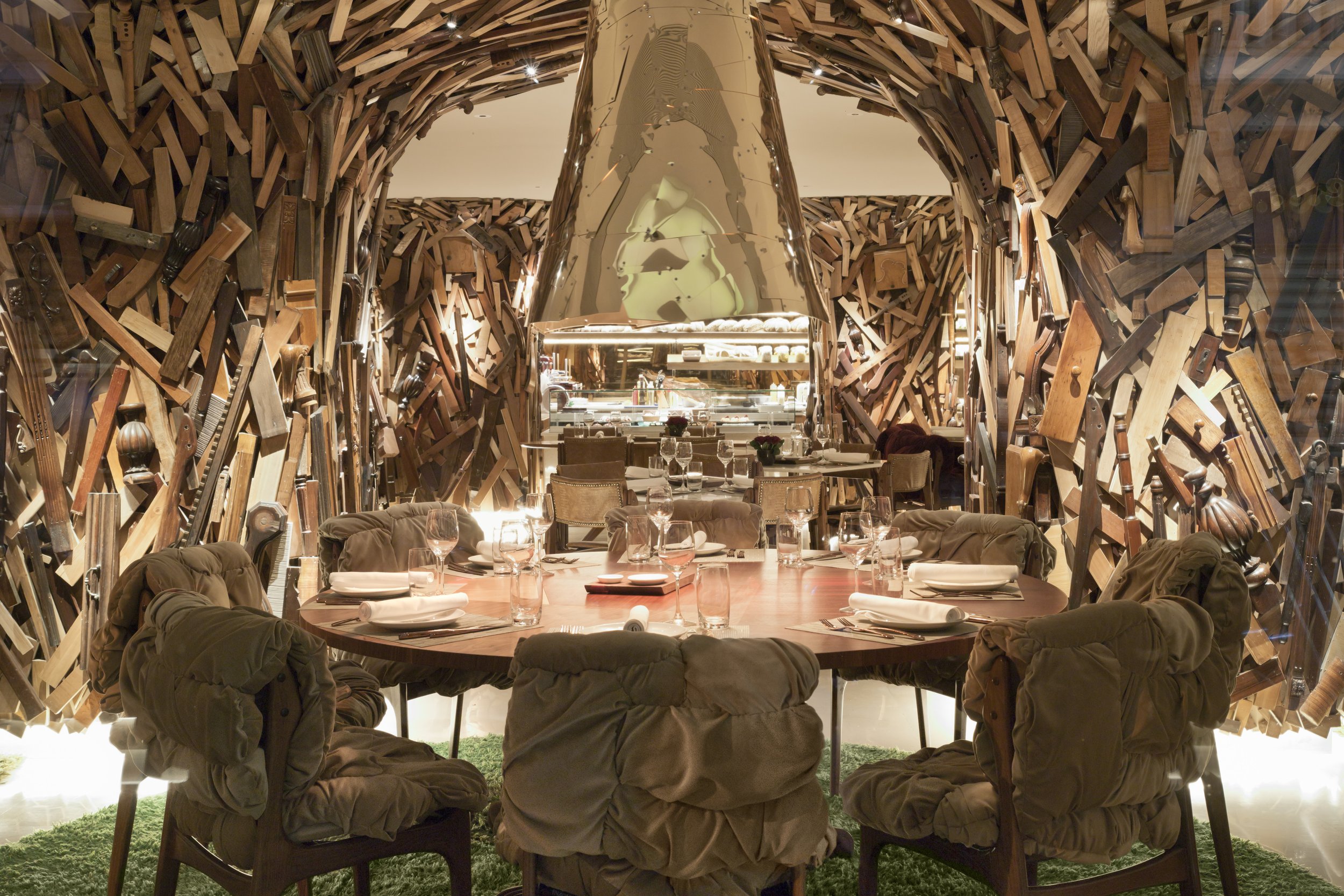 A restaurant with a unique interior featuring walls made of stacked, variously sized wood pieces. A round dining table is set with glasses, plates, and napkins, surrounded by cushioned chairs. NEW Hotel Athens Greece