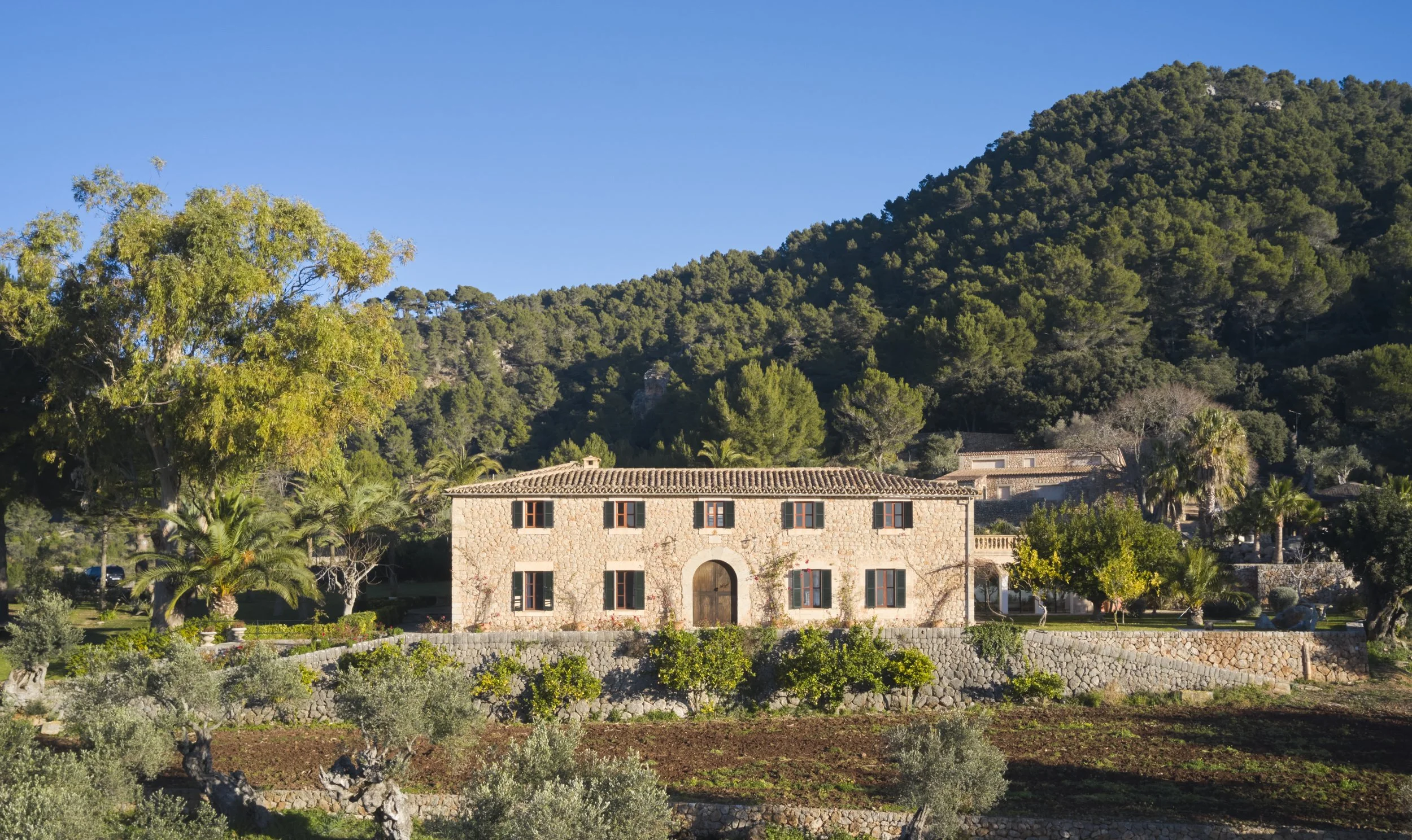 A stone house with red window shutters and a tiled roof sits amidst lush greenery and trees, with a backdrop of a hillside covered in dense forest under a clear blue sky. Vestige Collection Son Veri Mallorca Spain