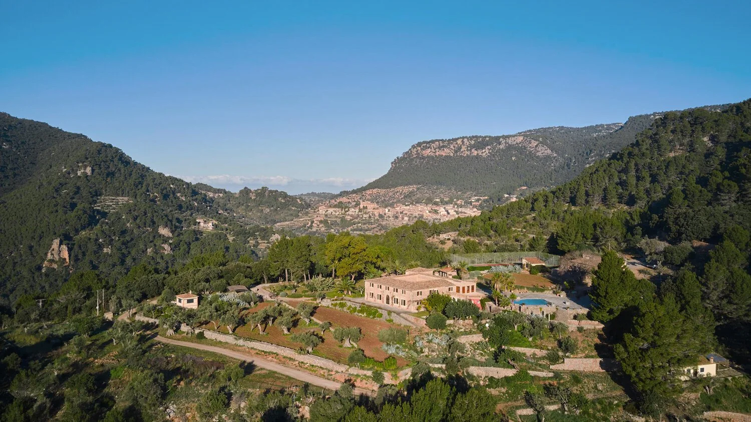 Aerial view of a hillside estate with a house, swimming pool, and terraced gardens surrounded by trees and mountains. Vestige Collection Son Veri Mallorca Spain