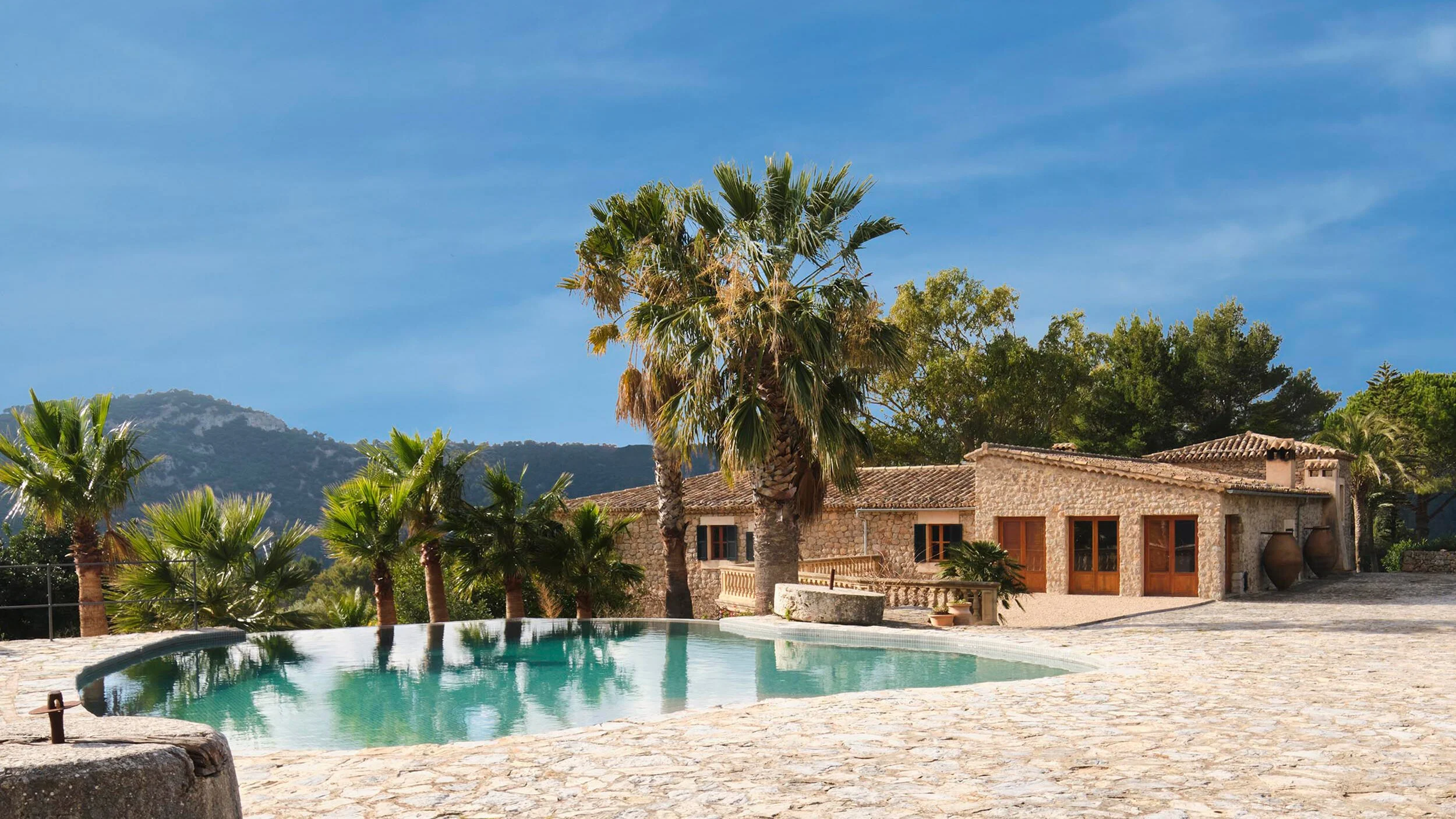 View of a stone house with wooden doors, a swimming pool, and palm trees against a mountain and blue sky background. Vestige Collection Son Veri Mallorca Spain