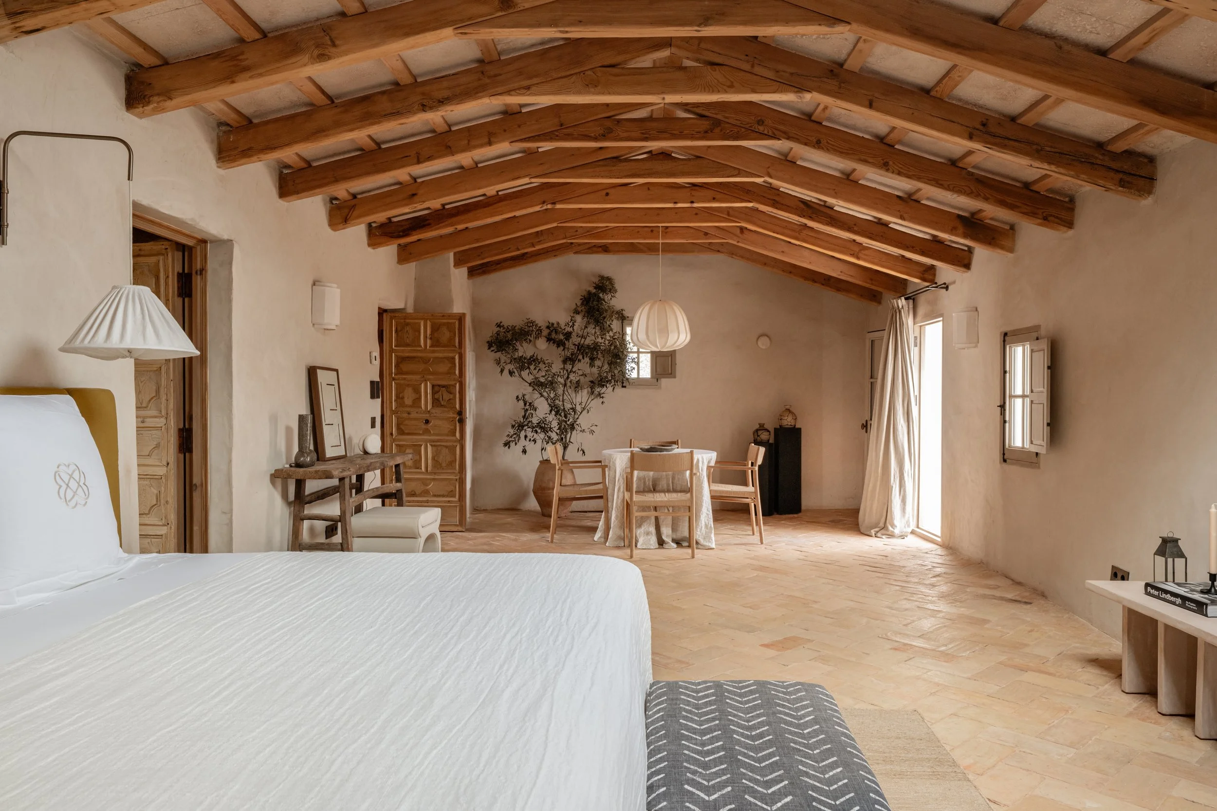 A spacious bedroom with a rustic design, featuring a white bed, wooden ceiling beams, a round dining table with four chairs, a large potted plant, and multiple windows with shutters and curtains. Vestige Collection Son Ermita Binidufa Menorca Spain
