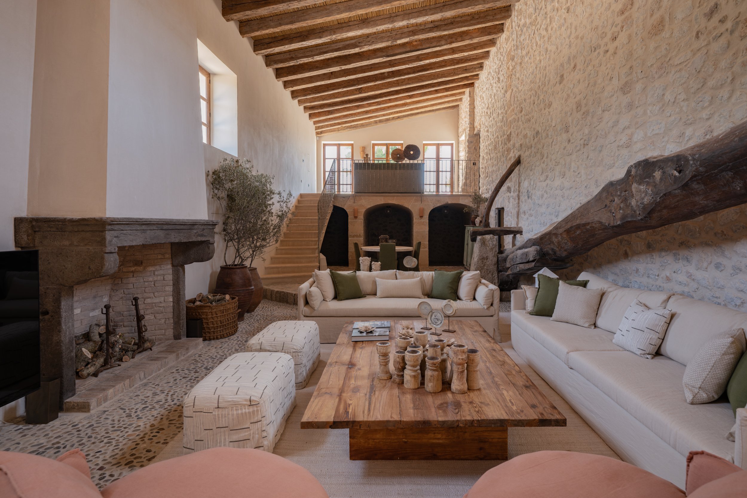 Vestige Son Veri Mallorca - Interior of a rustic living room featuring a stone and wood design with large windows, a fireplace, a white sofa with green pillows, a wooden coffee table, and decorative vases on a staircase landing.