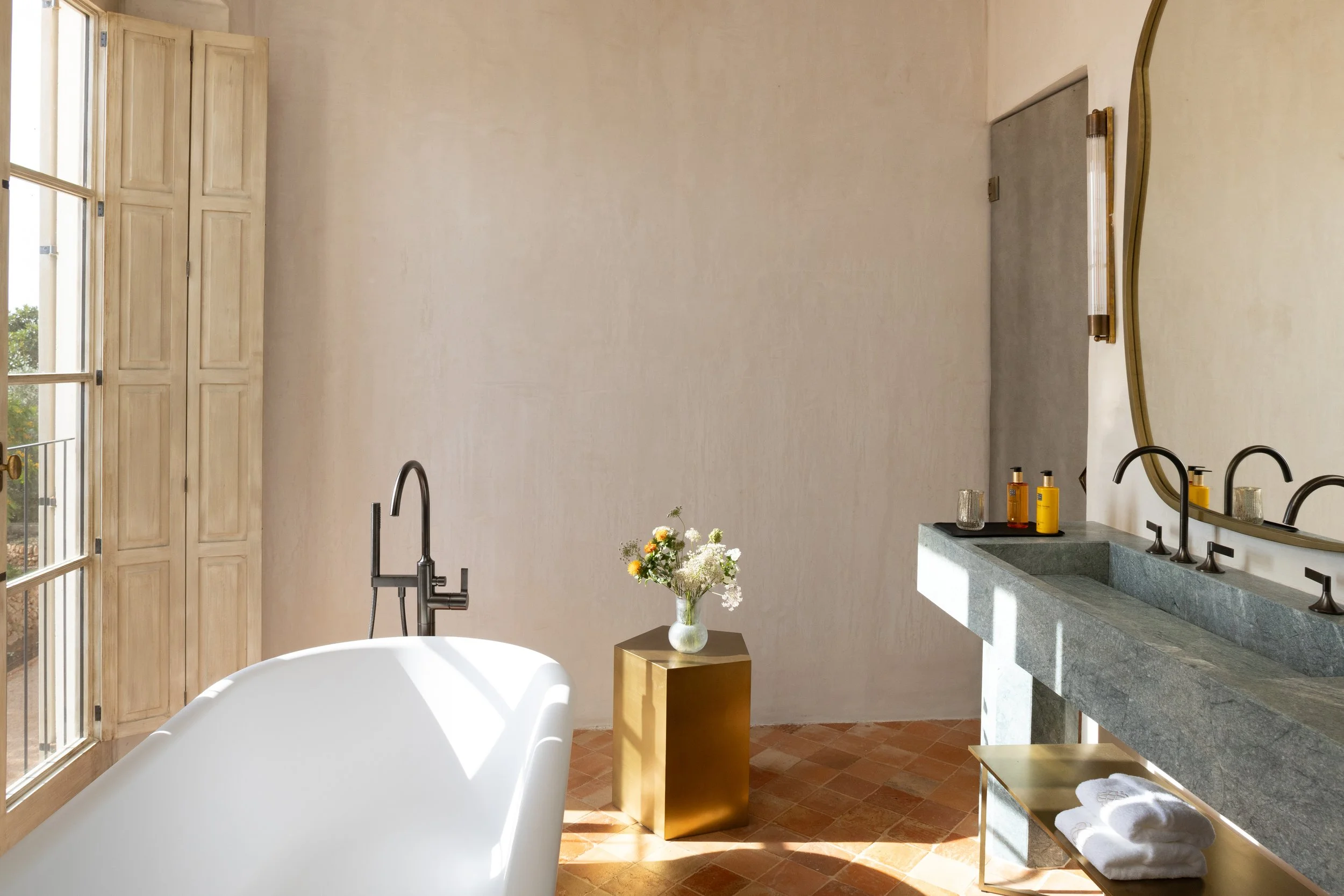Modern bathroom with a freestanding bathtub, a small golden side table with a flower vase, a green stone vanity with two sinks, a large oval mirror, decorative bottles, towels, wooden shutters. Vestige Collection Son Vell Menorca Spain