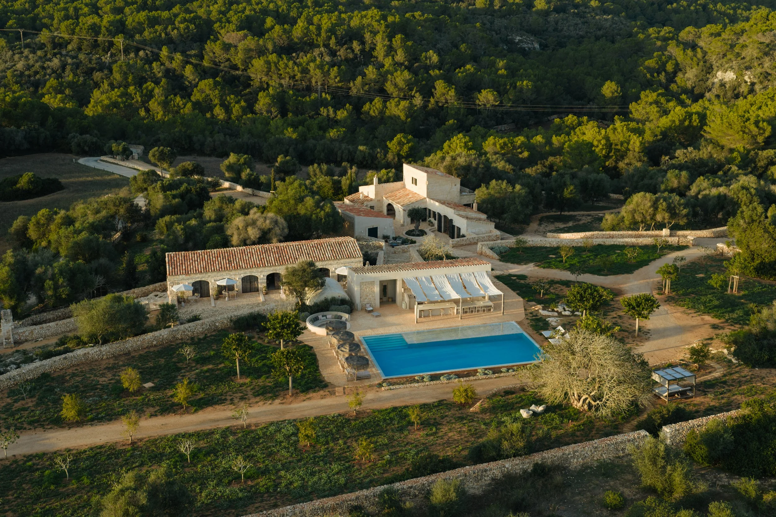 Aerial view of a Mediterranean-style estate with a swimming pool, surrounding greenery, and lush trees with a forest backdrop. Vestige Collection Santa Ana Menorca Spain