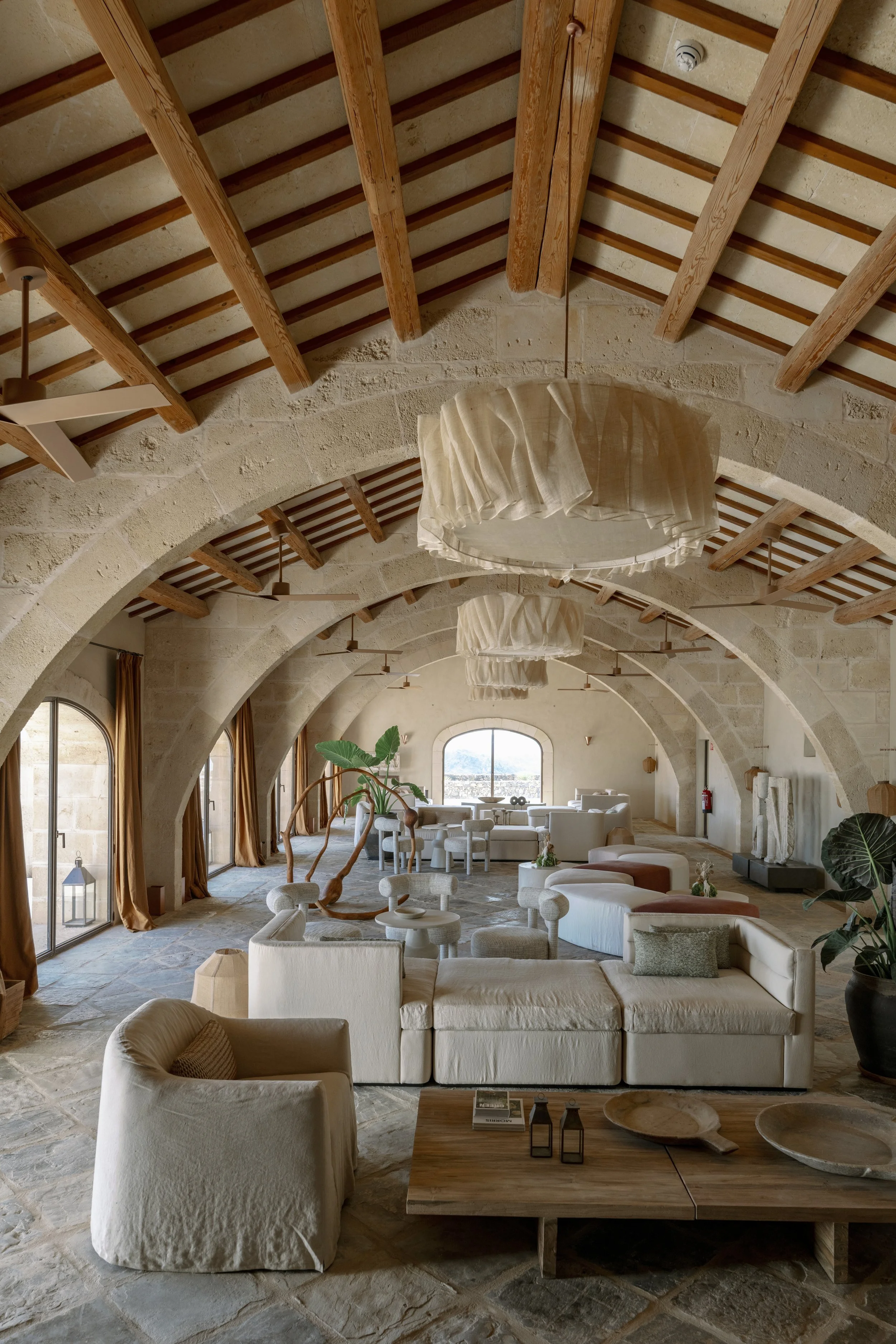 Interior of a spacious living room with arched stone walls, wooden ceiling beams, large vaulted windows, and modern beige furniture, including sofas and armchairs. Vestige Collection Son Ermita Binidufa Menorca Spain