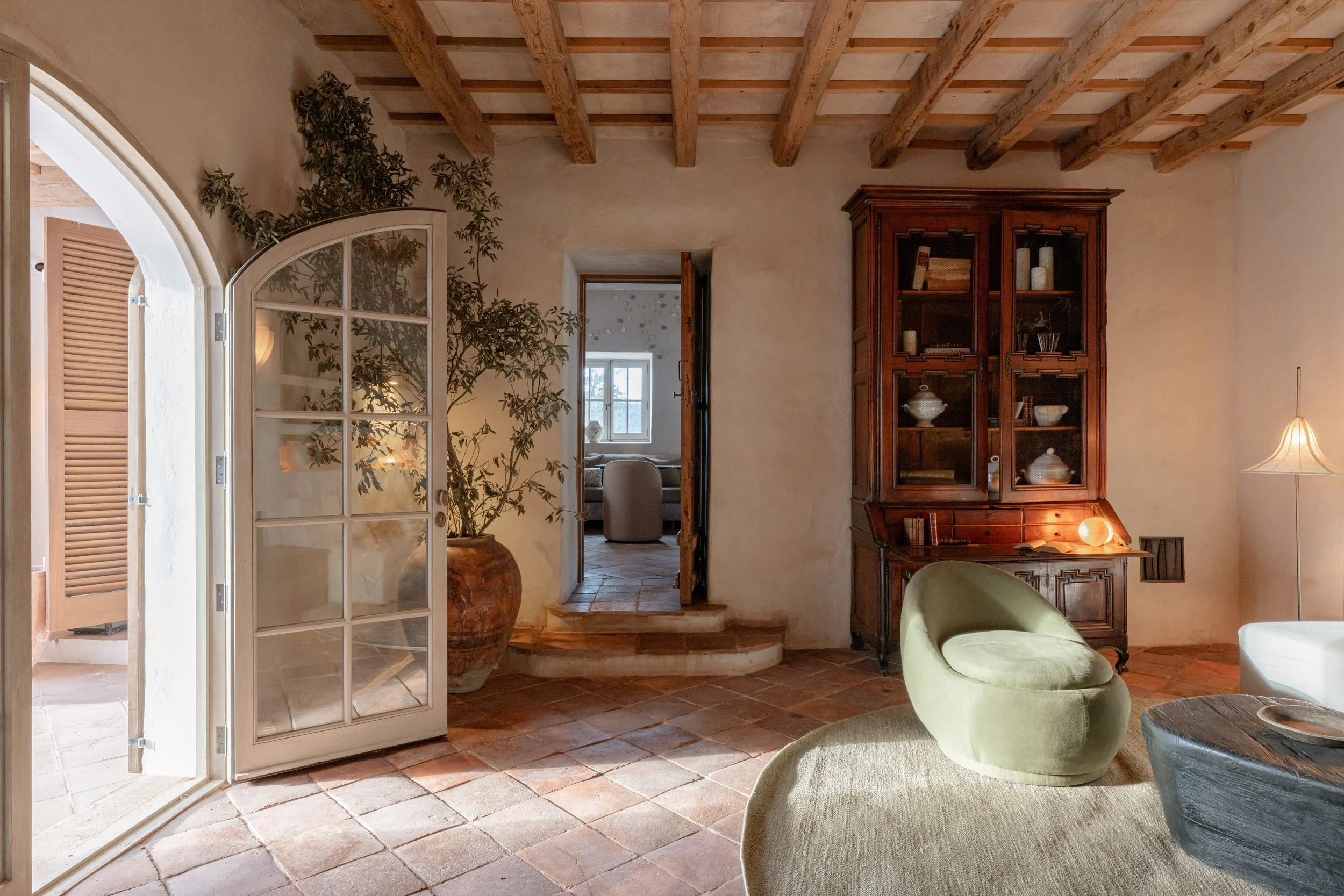 A cozy living room with terracotta tile flooring, a large potted plant near a glass-paned door, a wooden cabinet displaying dishes, and a modern green lounge chair. Vestige Collection Son Ermita Binidufa Menorca Spain