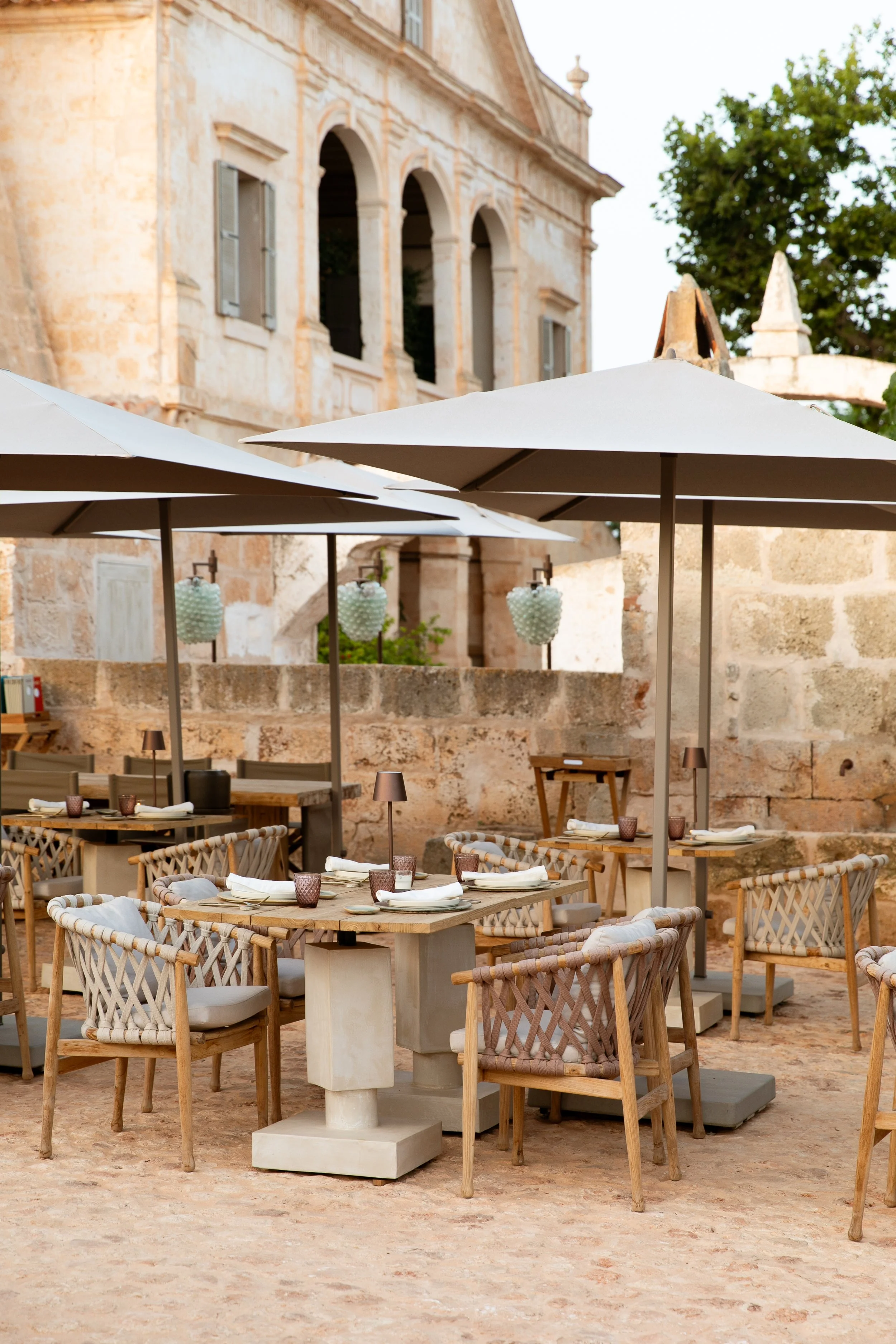 Outdoor restaurant seating area with wooden tables, woven chairs, white napkins, glasses, and small lamps under large umbrellas, with a historic stone building in the background. Vestige Collection Son Vell Menorca Spain