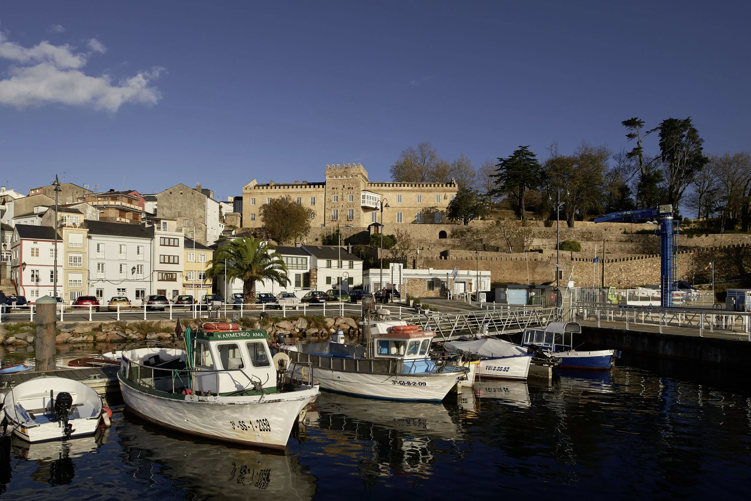 Boats docked at a harbor with colorful buildings, a castle, trees, and a blue sky in the background. Vestige Collection Palacio de Figueras Asturias Spain