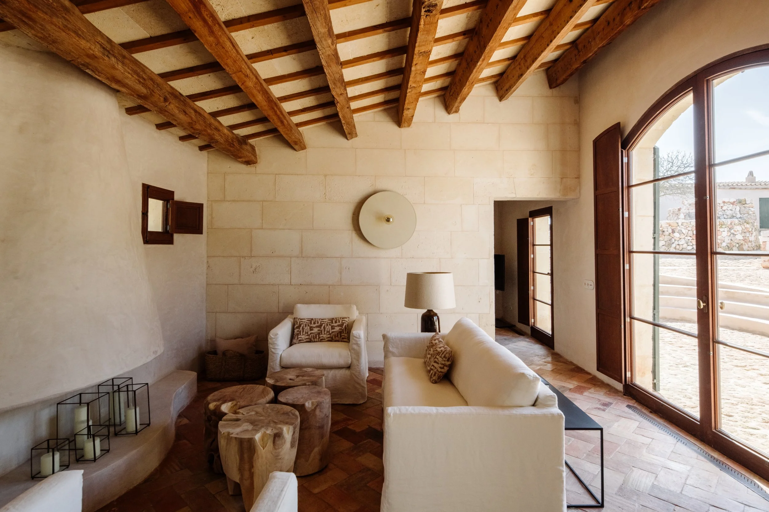 Living room with cream-colored sofas, wooden coffee tables, a floor lamp, wooden ceiling beams, beige stone walls, a large arched window, and small windows on the side walls. Vestige Collection Santa Ana Menorca Spain