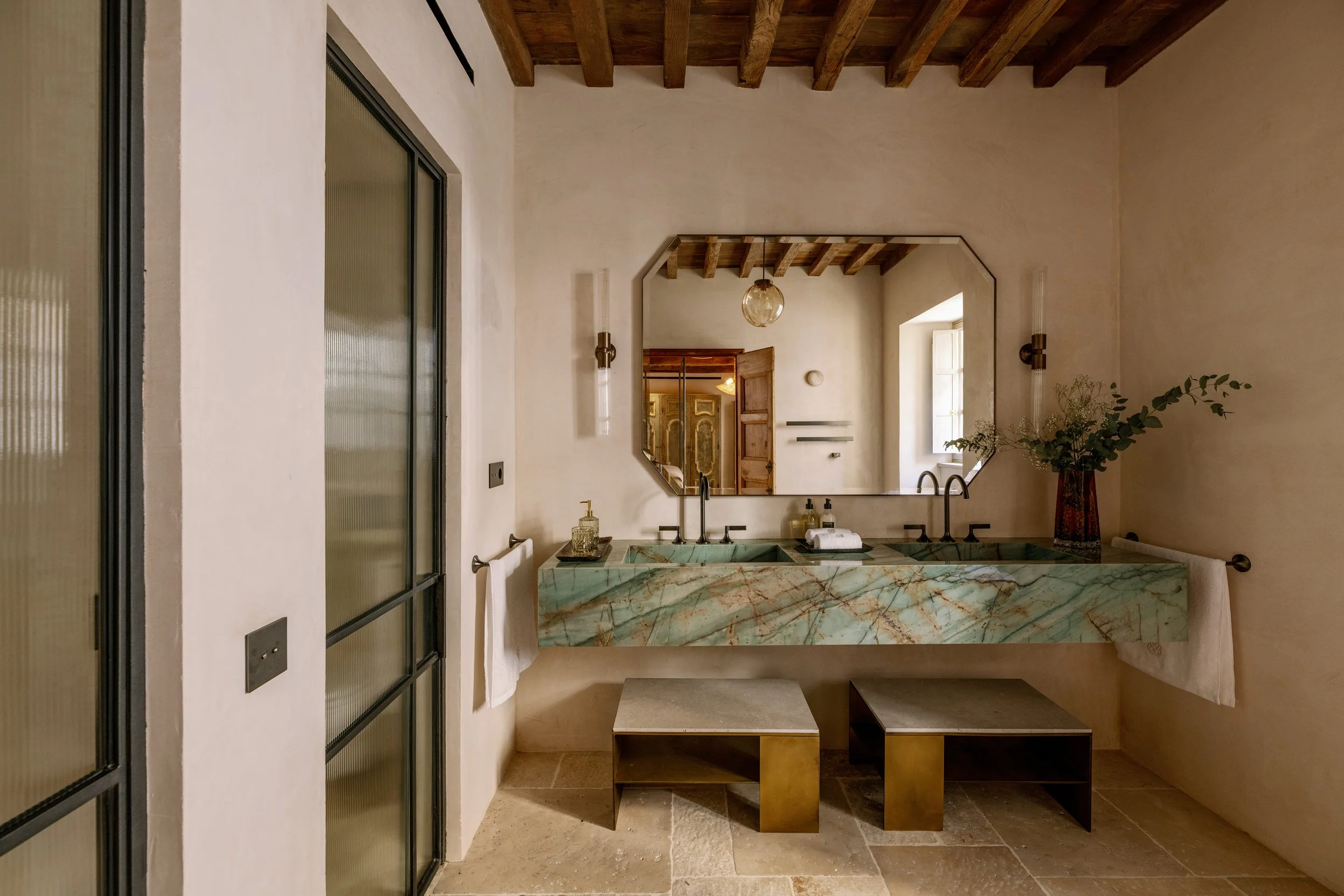 Bathroom with a double green marble sink, large mirror, and potted plant, with a wooden beamed ceiling and natural light from a window. Vestige Collection Miramar Mallorca Spain