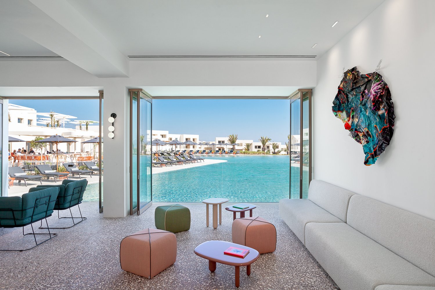 Modern interior with a large open window overlooking a swimming pool and resort outside, decorated with colorful seating, a long white sofa, and wall art. NOUS Santorini Hotel Greece