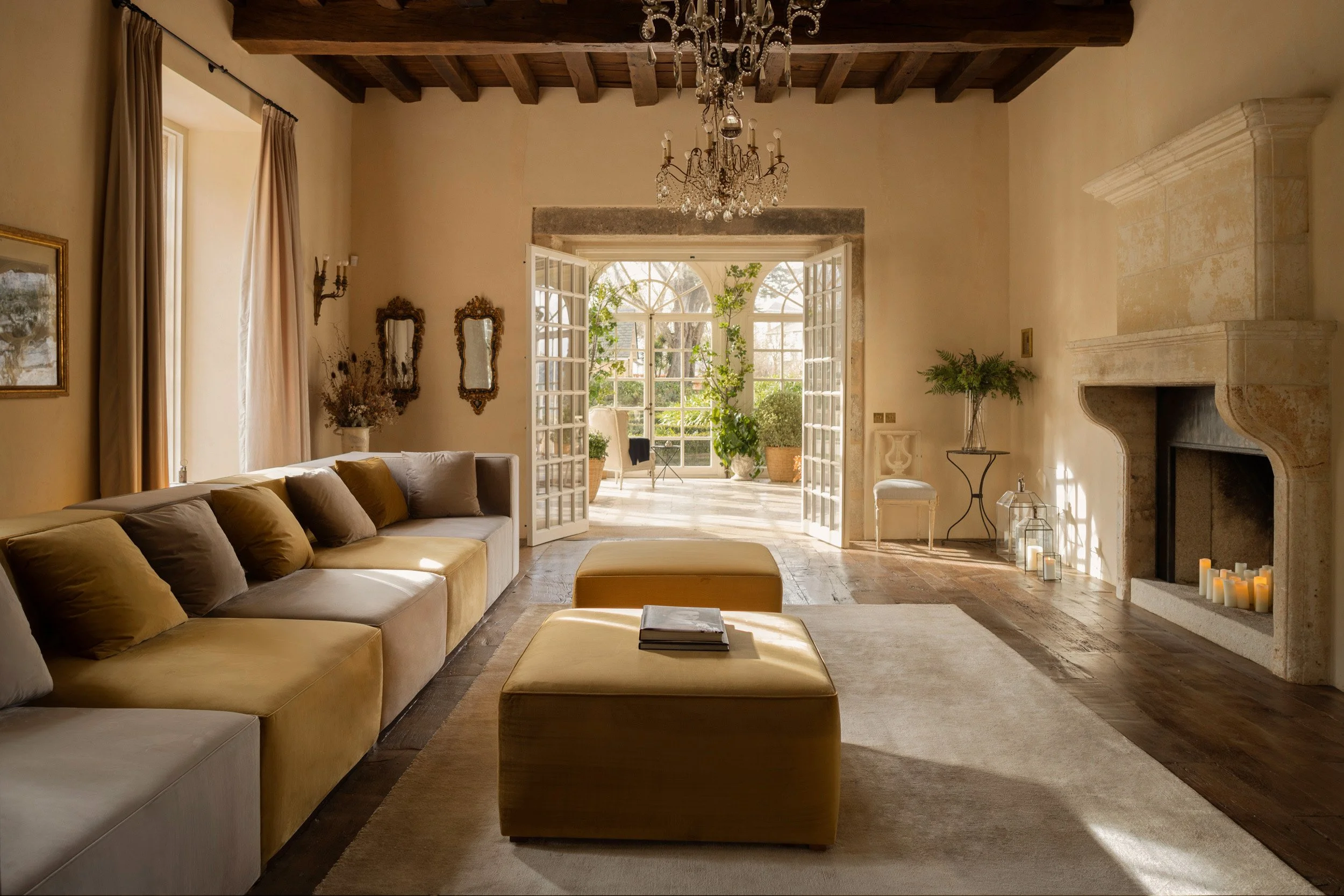 Elegant living room with a beige sofa, a yellow ottoman, and a fireplace with candles. Sunlit sunroom with potted plants is visible through open French doors. Vestige Collection Palacio de Figueras Asturias Spain