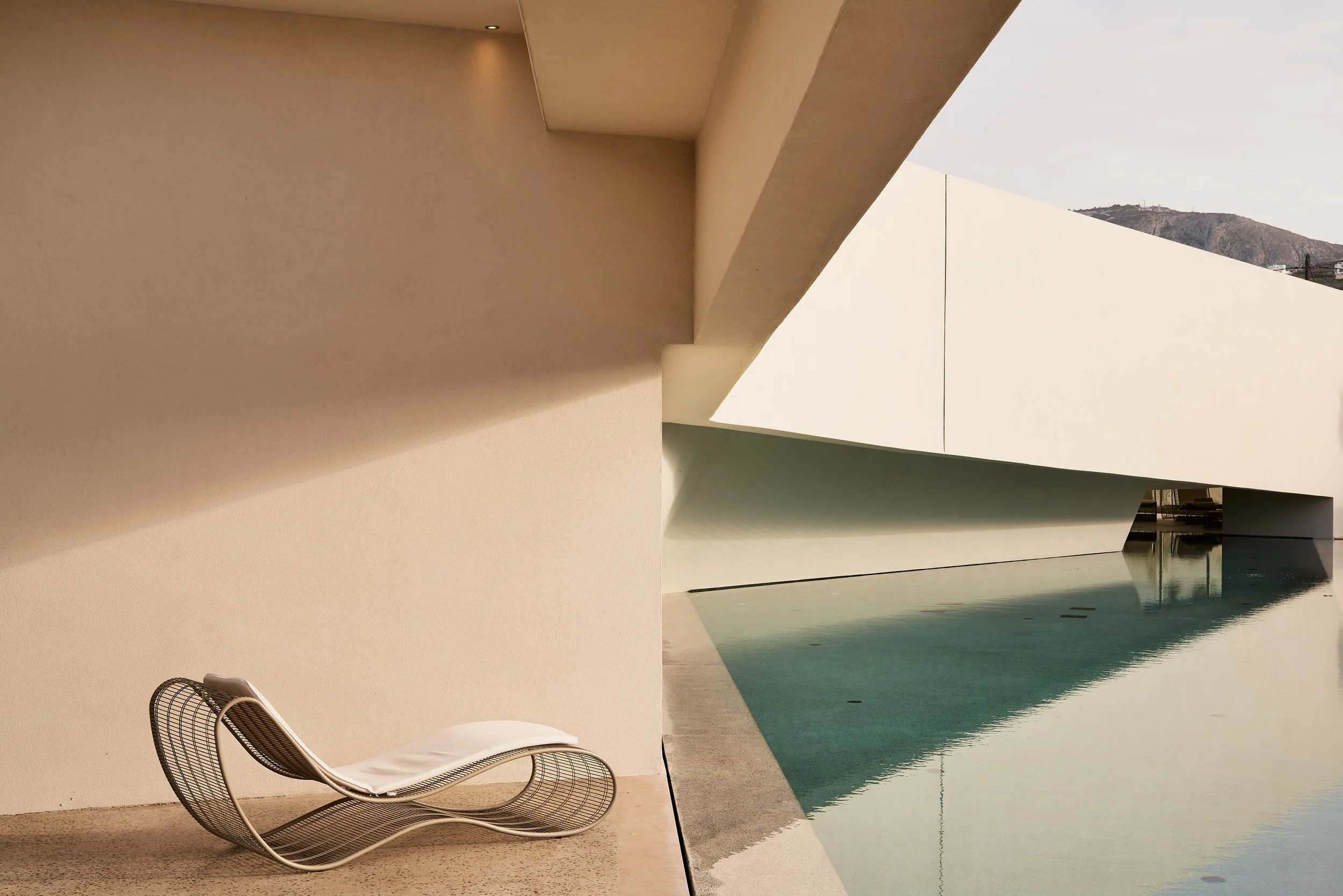 Modern minimalist building with geometric shapes and a reflective pool, featuring a lounge chair on a beige floor. NOUS Santorini Hotel Greece
