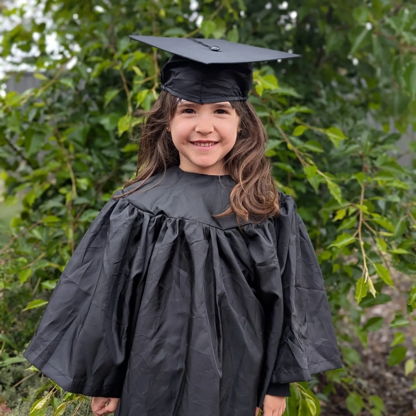Young girl in a black graduation gown and cap standing outdoors with green leafy background. Seeds Montessori child graduation from their class. End of Year ceremony.