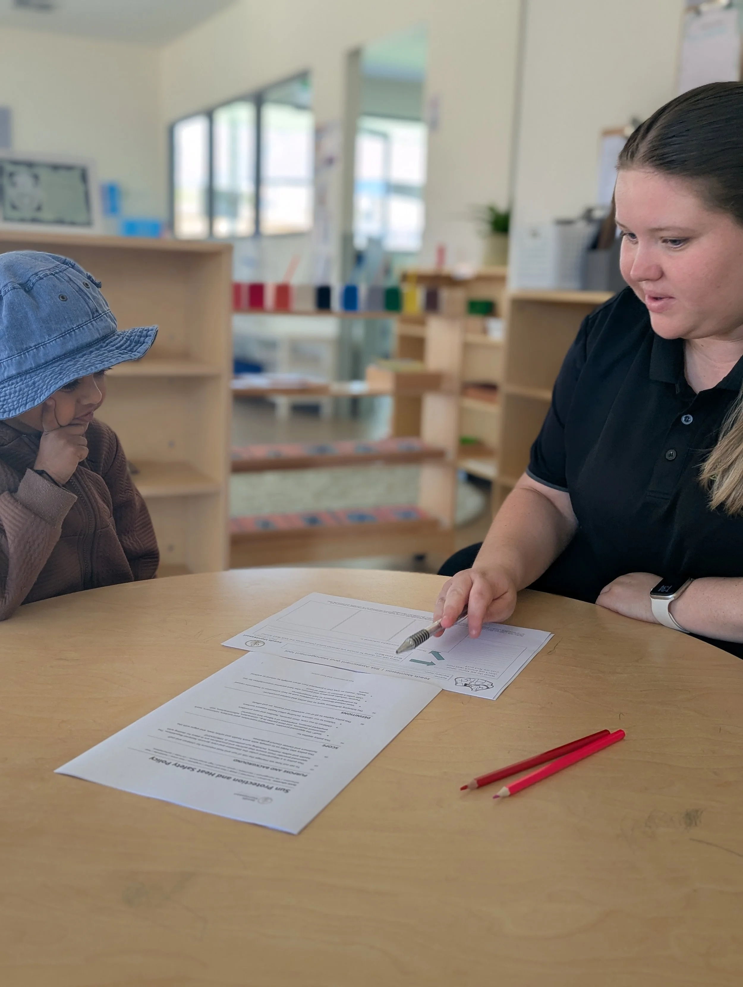 An Educator from Seeds Montessori, who is sitting at a wooden table in a classroom, handing a pen to a young girl wearing a blue hat. There are papers and red pencils on the table. The background shows learning materials, windows, and shelves.