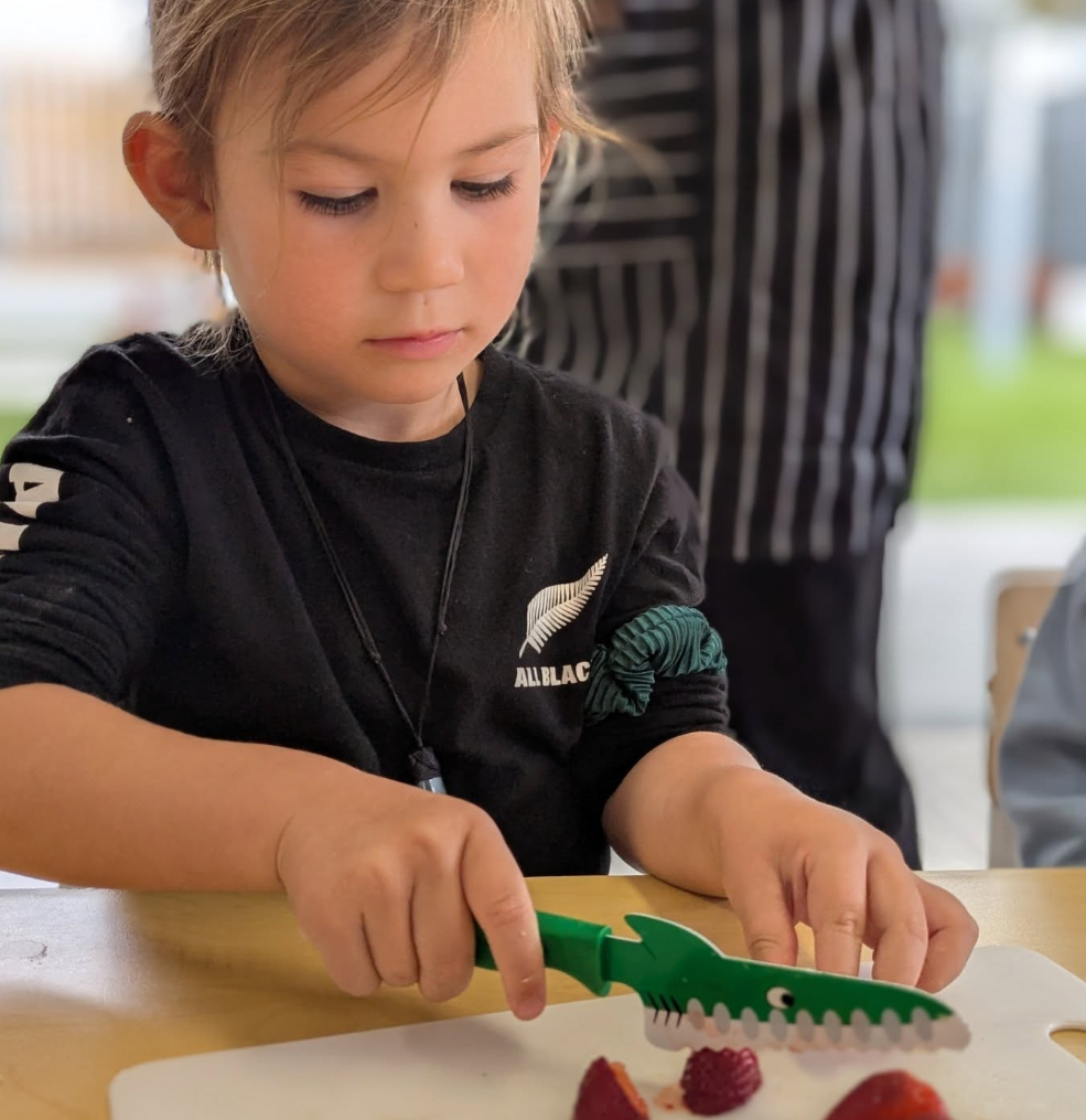 A young girl with blonde hair wearing a black shirt with a silver fern and the text 'ALL BLACK' is cutting a piece of cake or pastry on a white cutting board with a green dinosaur-shaped plastic knife. There are strawberries on the cutting board.