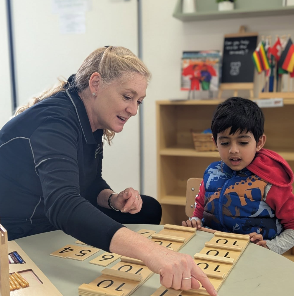 A woman teaching a young boy using wooden number tiles at a table in a classroom.
