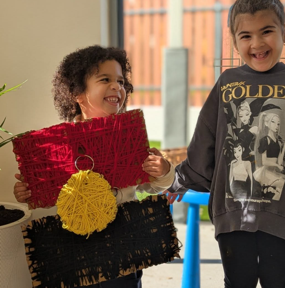 Two children holding colorful string art decorations, smiling and laughing.