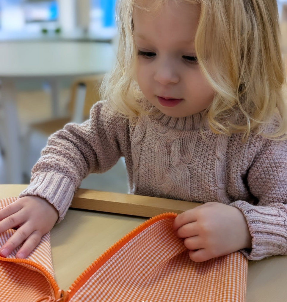 A young girl with blonde hair, wearing a pink knit sweater, at a table with an orange checkered bag, inside a bright room with blurred background.