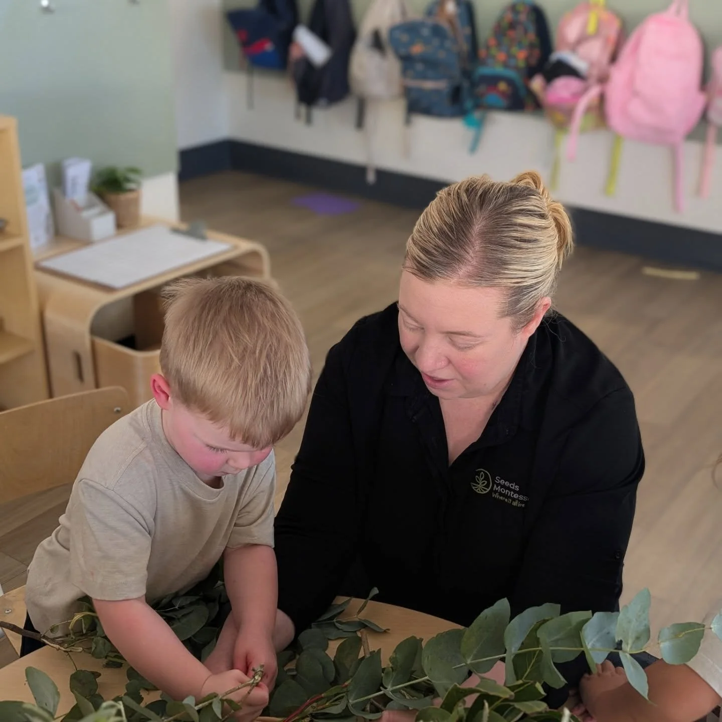 A young boy and an adult woman engaging in a hands-on activity with a branch of green leaves in a classroom setting. Centre Manager engaging with Seeds Montessori Child.