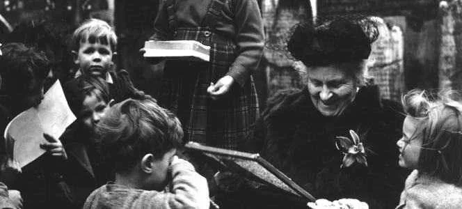 A woman surrounded by children outdoors, holding an open book or scrapbook, with children attentively looking at her.