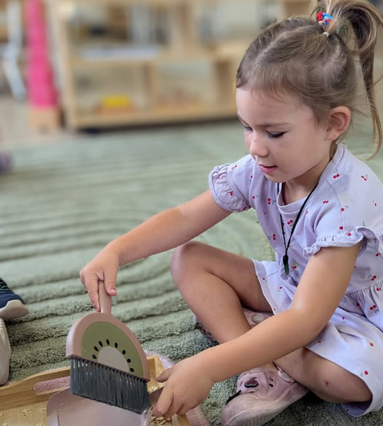 Young girl sitting cross-legged on a carpet, playing with a wooden toy that has a spinning wheel, in a room with shelves in the background.