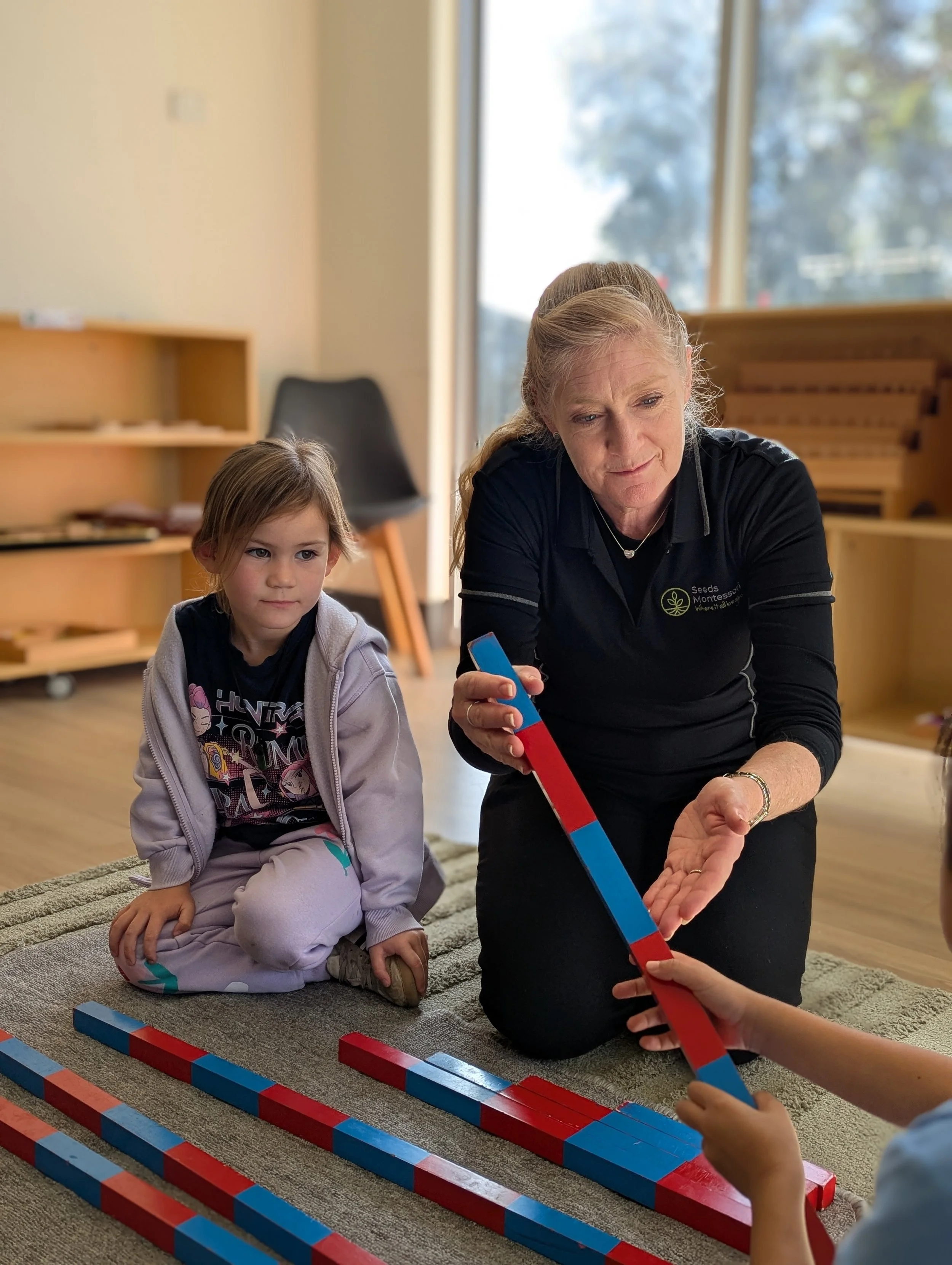 A woman and a young girl sitting on the floor playing with colorful, striped wooden blocks in a room with large windows.