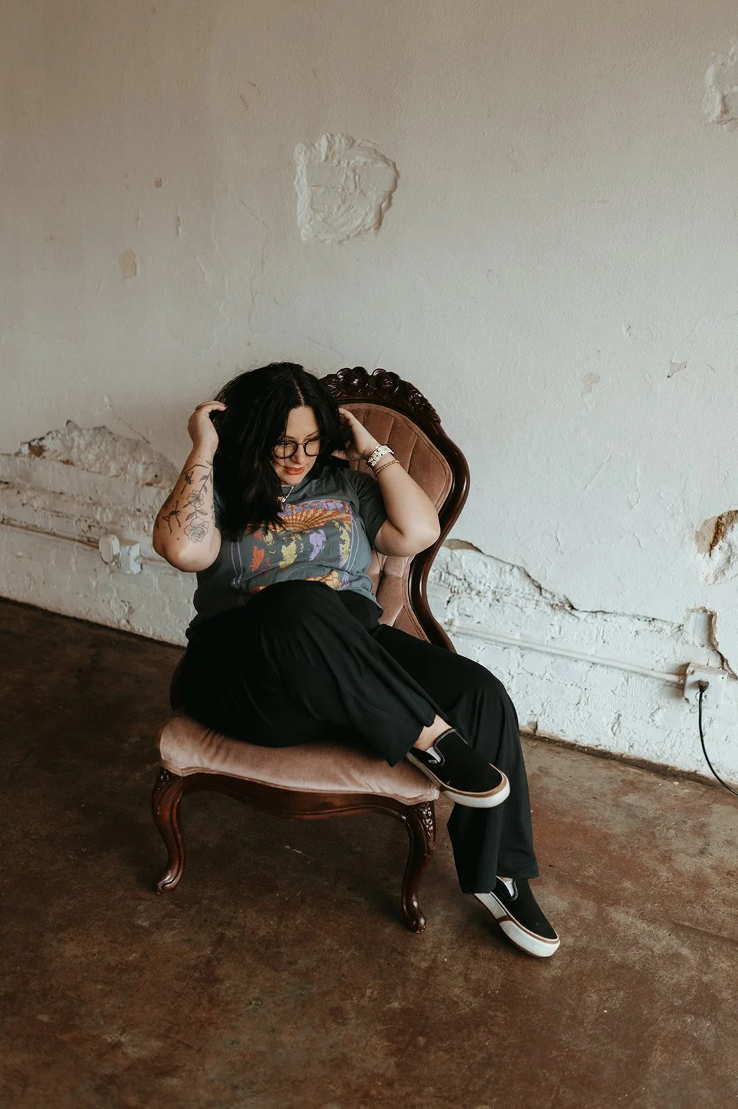 Cait McKeon with dark hair and glasses sitting on a vintage pink upholstered chair with a carved wooden frame, in front of a distressed white wall, wearing a graphic t-shirt, black pants, and slip-on sneakers, adjusting her hair.