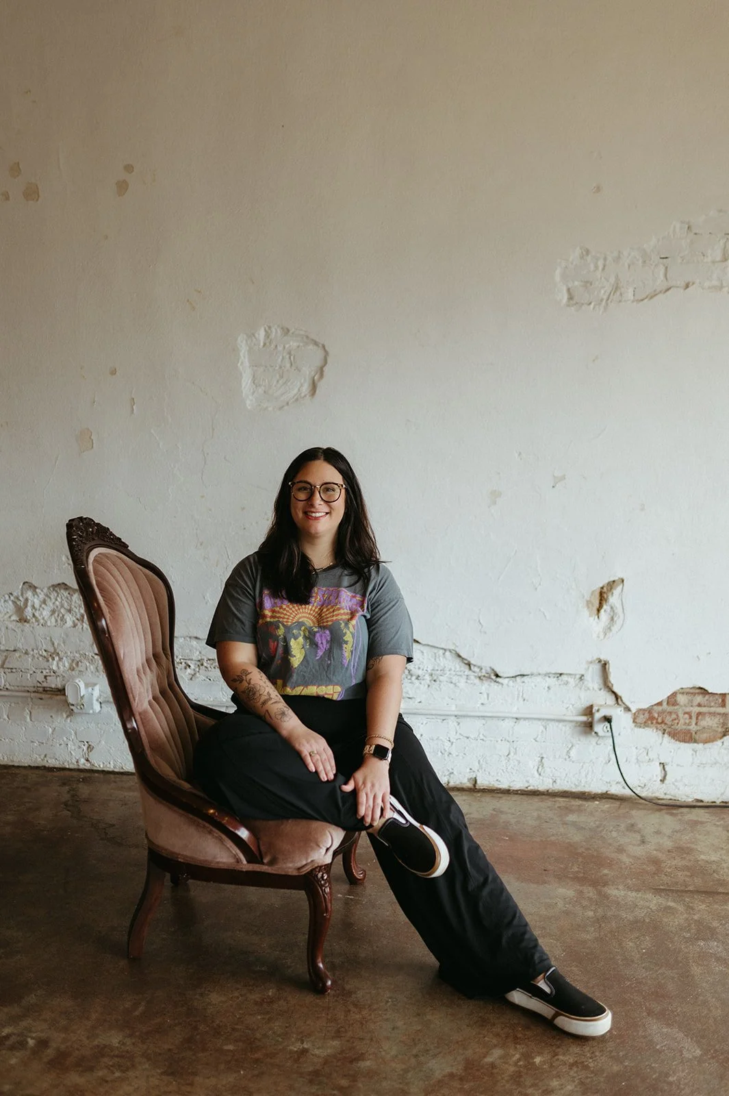 Cait McKeon with dark hair, glasses, and tattoos is sitting on a vintage upholstered armchair with wooden frame in an industrial-style room with a white brick and plaster wall, concrete floor, and visible electrical outlets.