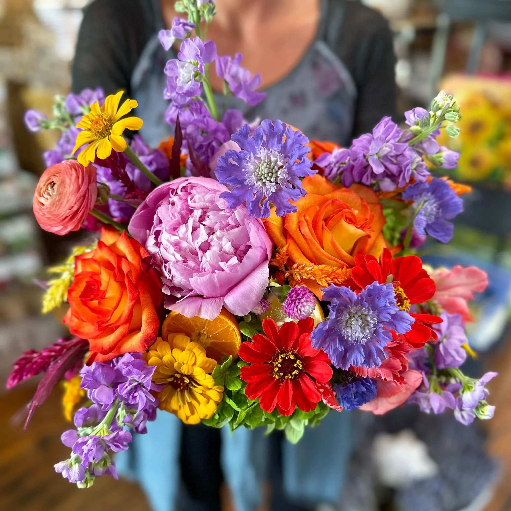 A colorful bouquet of various flowers, including purple, pink, orange, yellow, and red blooms, held by a person wearing a gray and black shirt in an indoor setting.