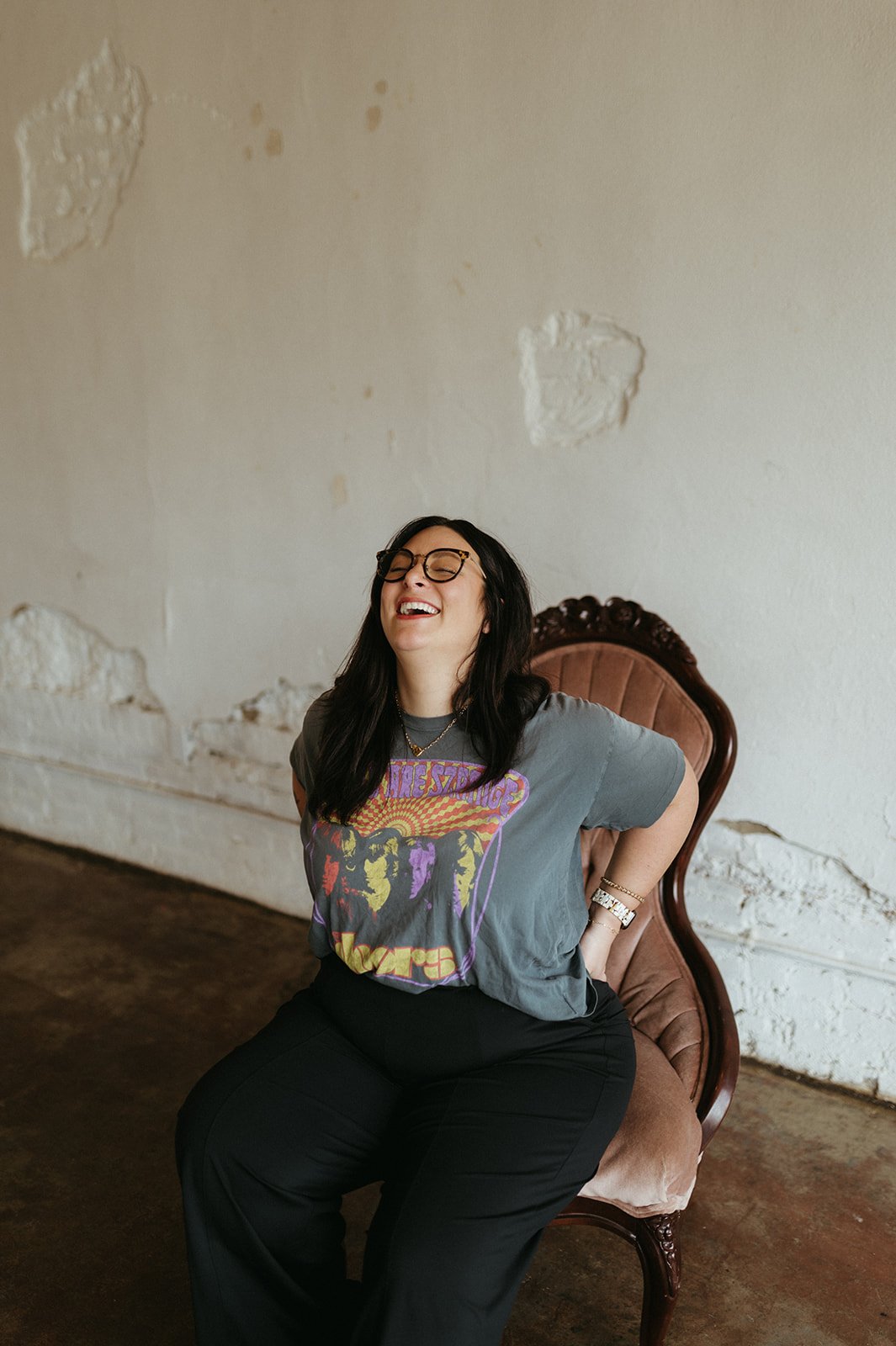 cait mckeon with glasses, long dark hair, wearing a graphic t-shirt, black pants, and accessories, sitting on a vintage upholstered chair with a carved wooden frame, laughing with her head tilted back.
