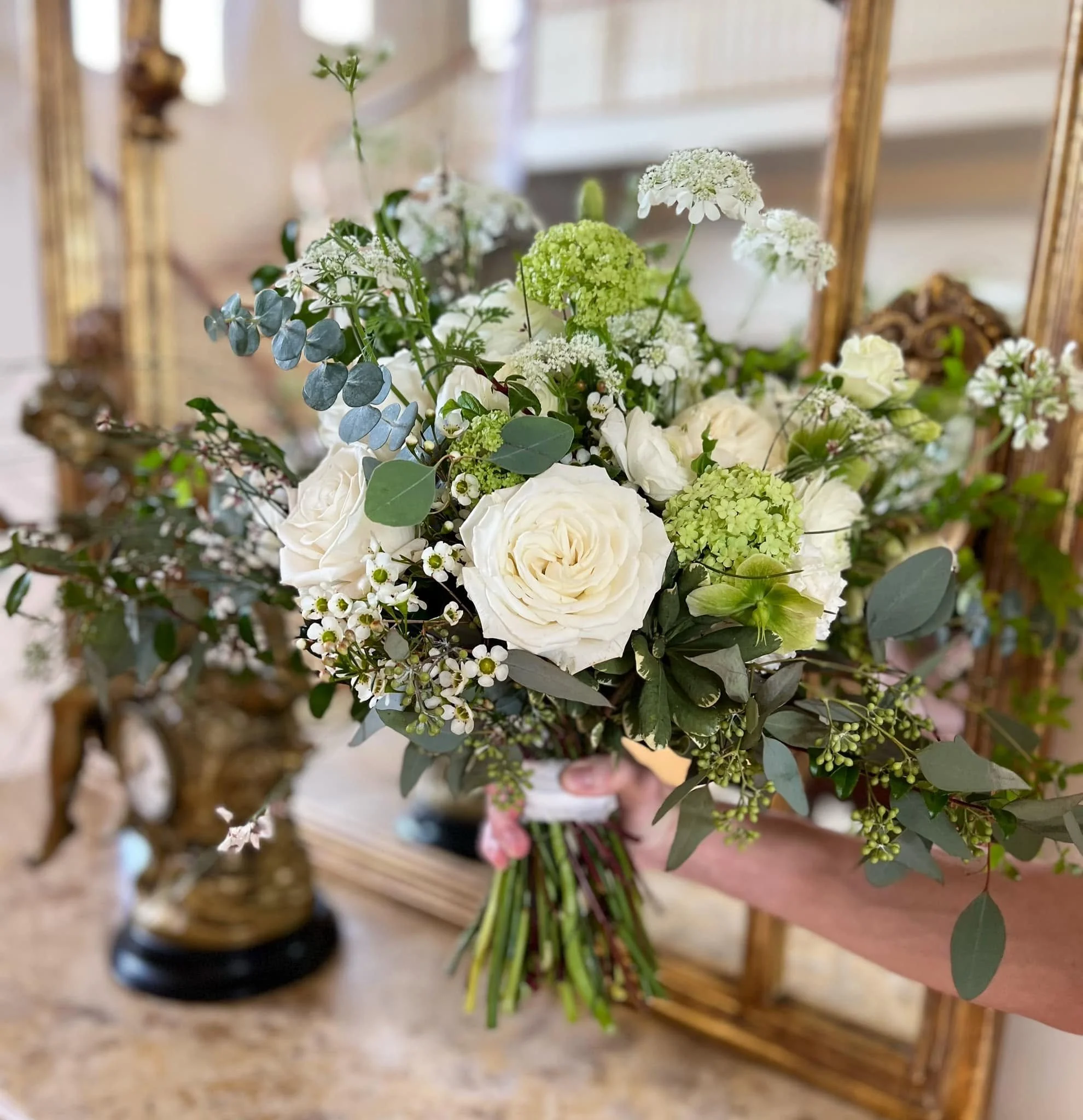 Close-up of a bouquet of white and green flowers, including roses and hydrangeas, held in a person's hand in front of a mirror with a gold frame.