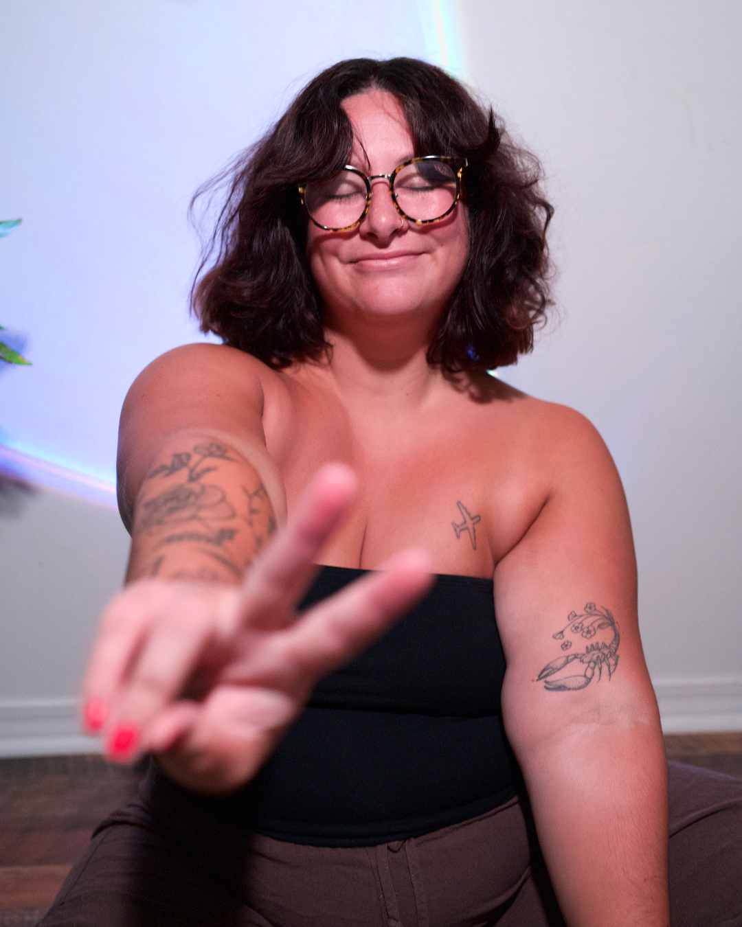 A woman with curly dark hair, wearing glasses, making a peace sign with her fingers, smiling with her eyes closed, and tattoos on her arm and chest.