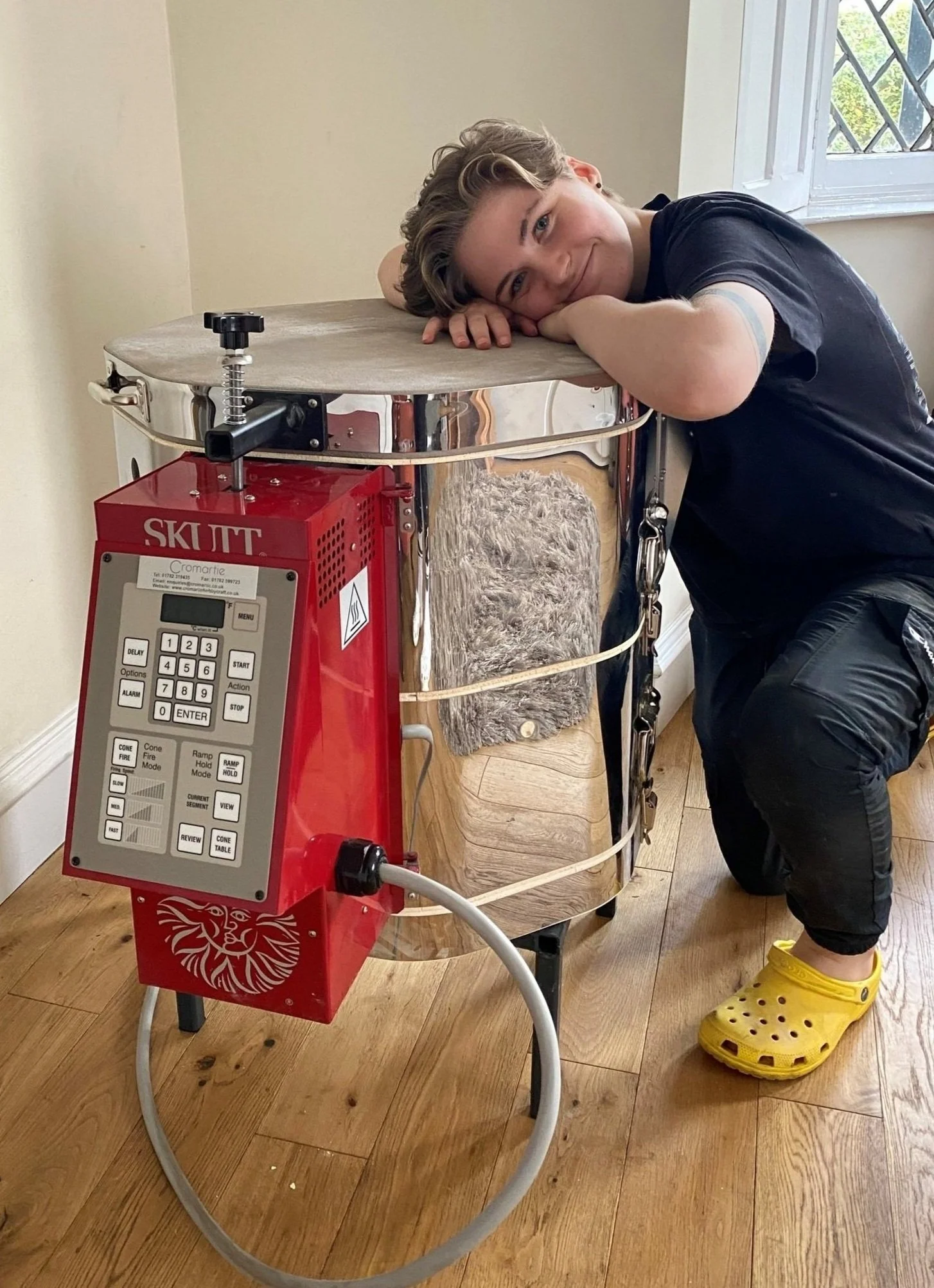 Young person resting their head on a kiln, smiling, wearing yellow Crocs, with a furry mat on the metal surface in a room with wooden flooring and a window.