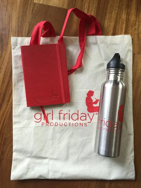 A cream tote bag, silver water bottle, and red journal all display the Girl Friday Productions logo