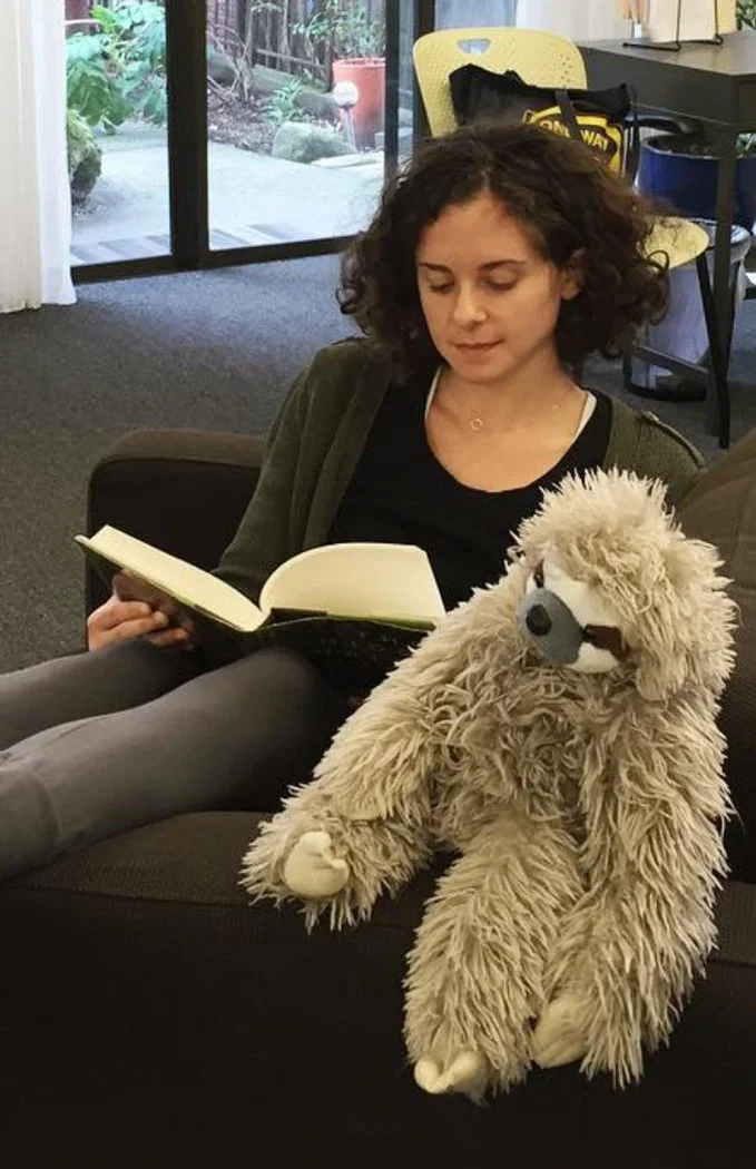 A young woman with curly hair reads a book in a chair with a sloth stuffy on the arm