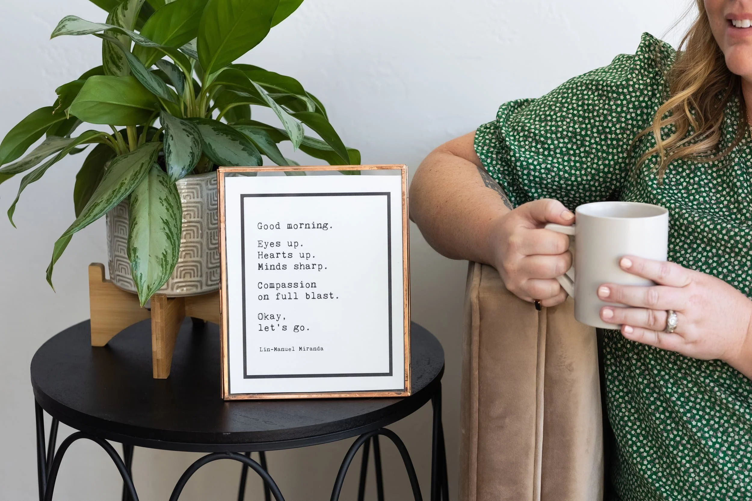 A woman sitting on a beige chair holding a white mug in her right hand, wearing a green patterned dress. On a small black table next to her, there is a potted plant with green leaves and a framed quote that reads: "Good morning. Eyes up. Hearts up. Minds sharp. Compassion on full blast. Okay, let's go."