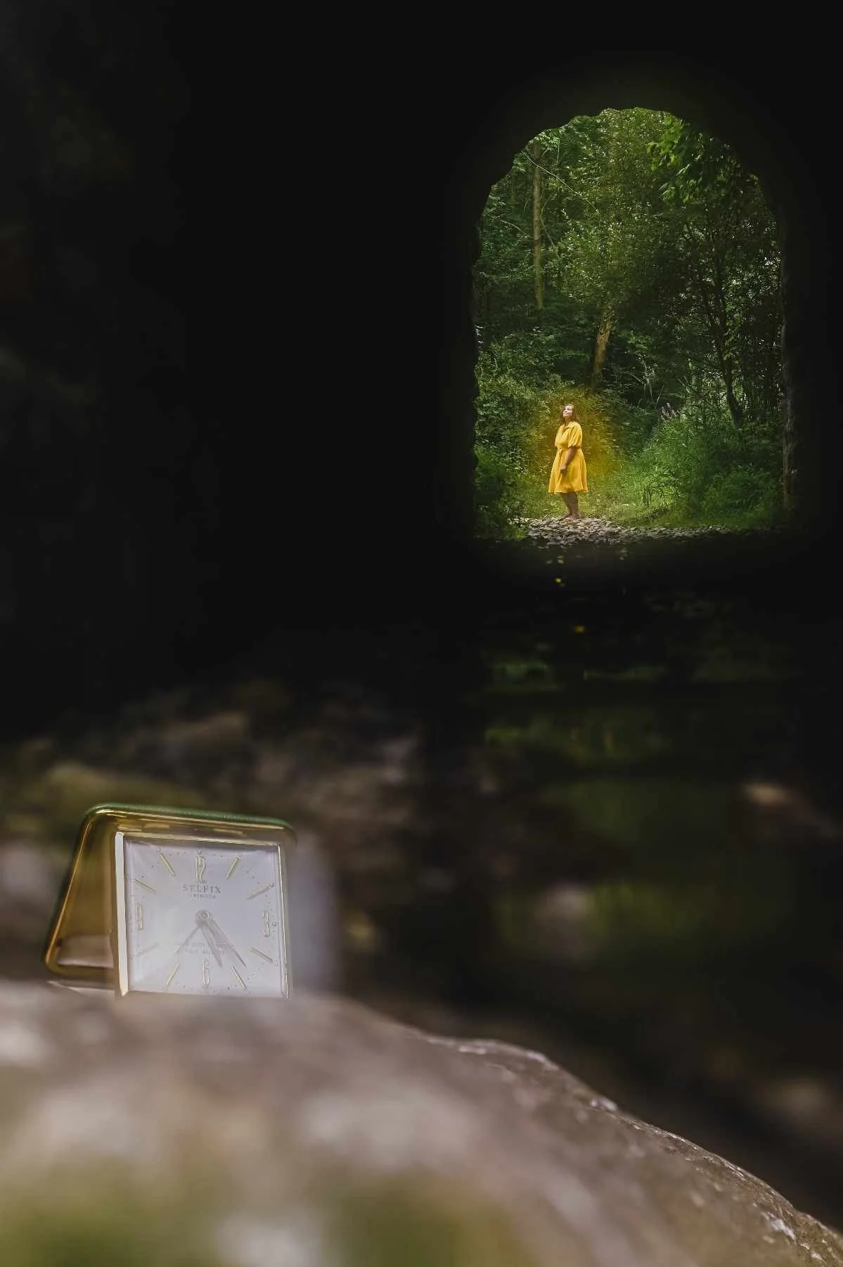 View through a tunnel showing a woman in a yellow coat standing in a green forest, with a small clock on the ground in the foreground.