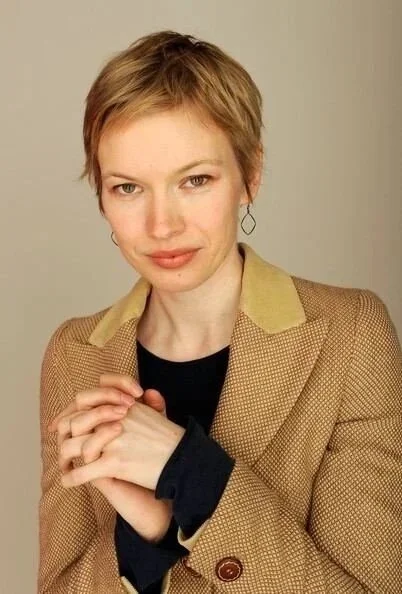 A woman with short, light brown hair, wearing a tan blazer over a black top, holding her hands together, looking at the camera with a slight smile.