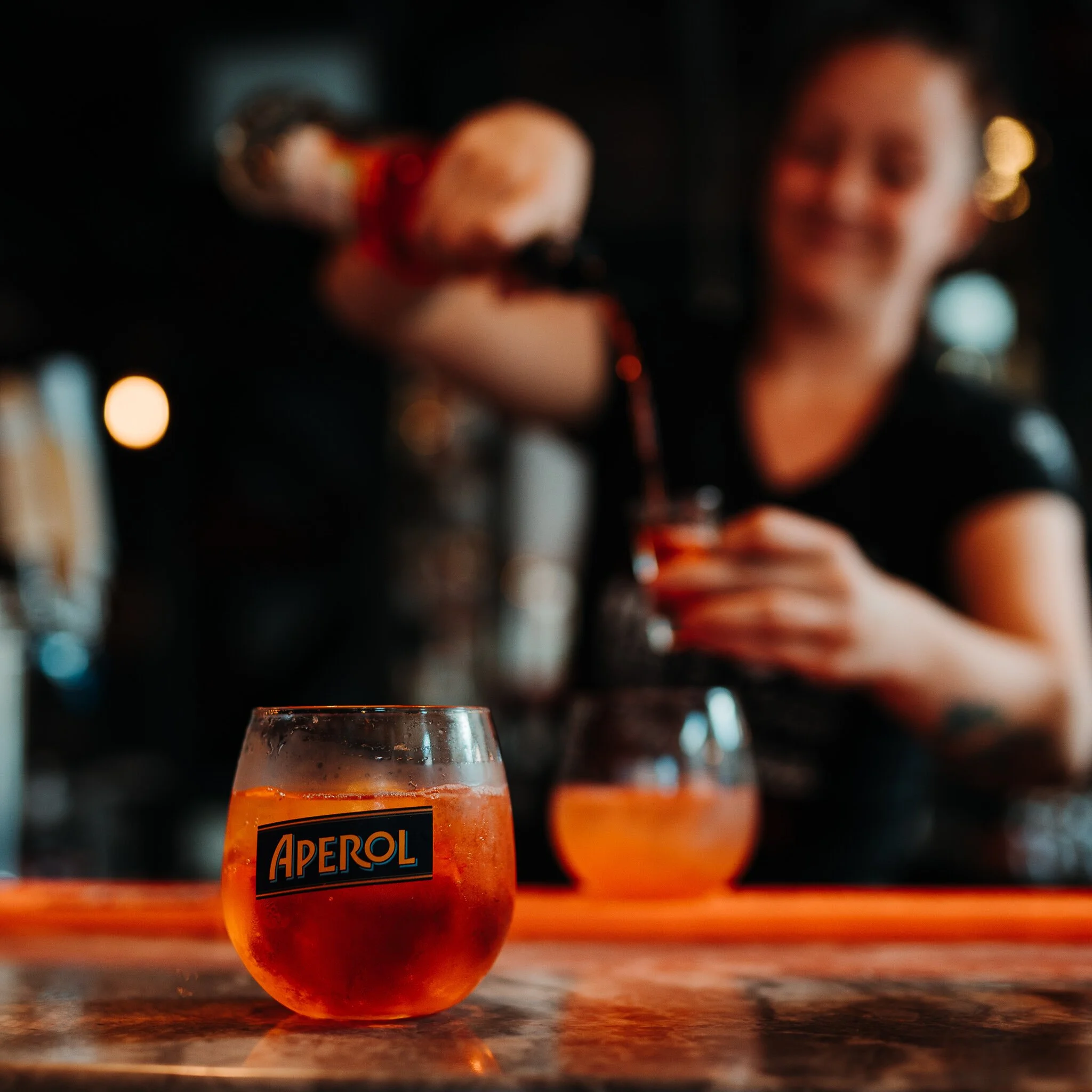 A glass of Aperol spritz on a bar counter with a bartender pouring more drinks in the background.