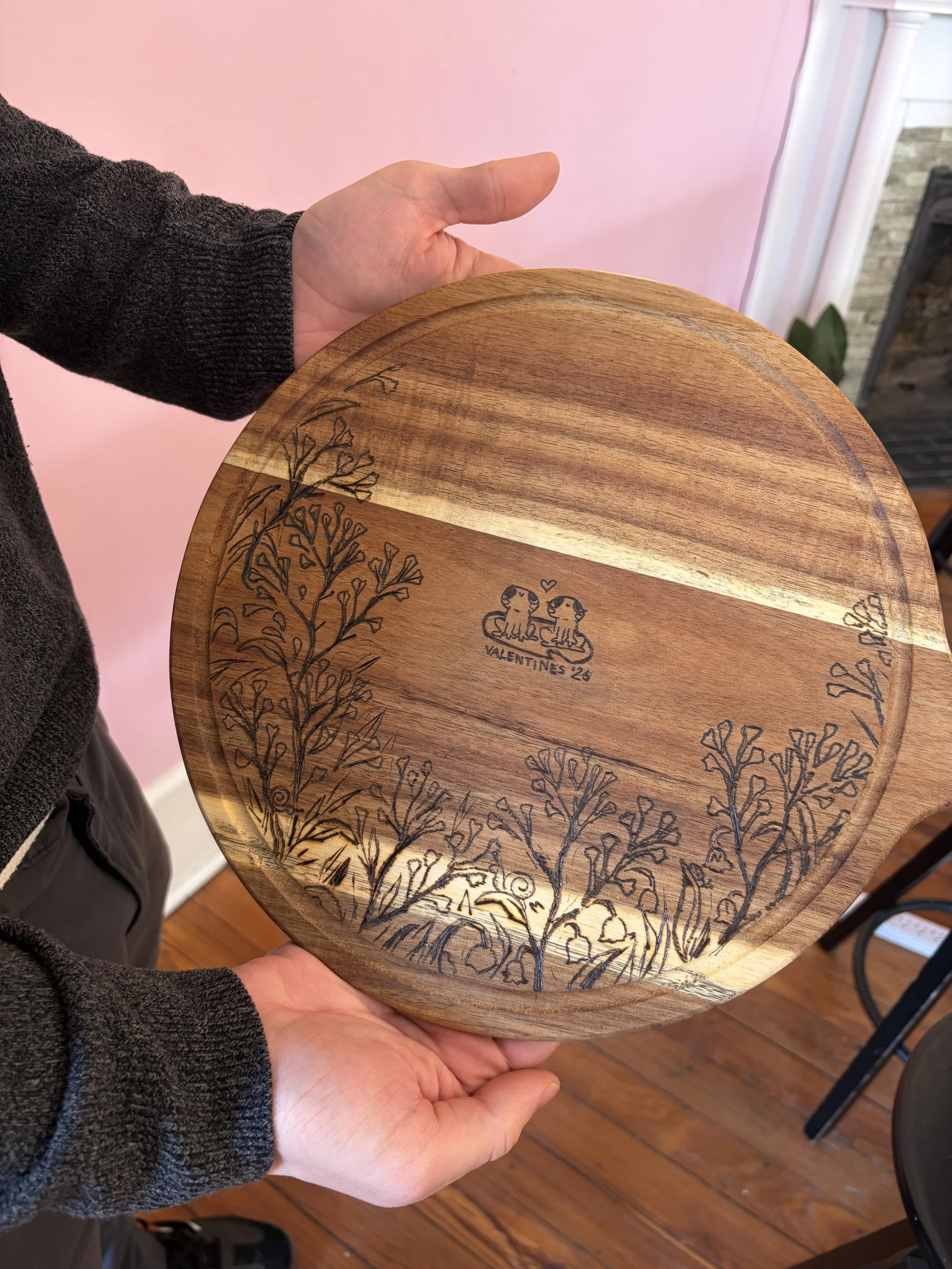 A person holding a wooden plate with engraved trees and two dogs with a heart between them. The plate has a note that says 'Valentine's 26'.