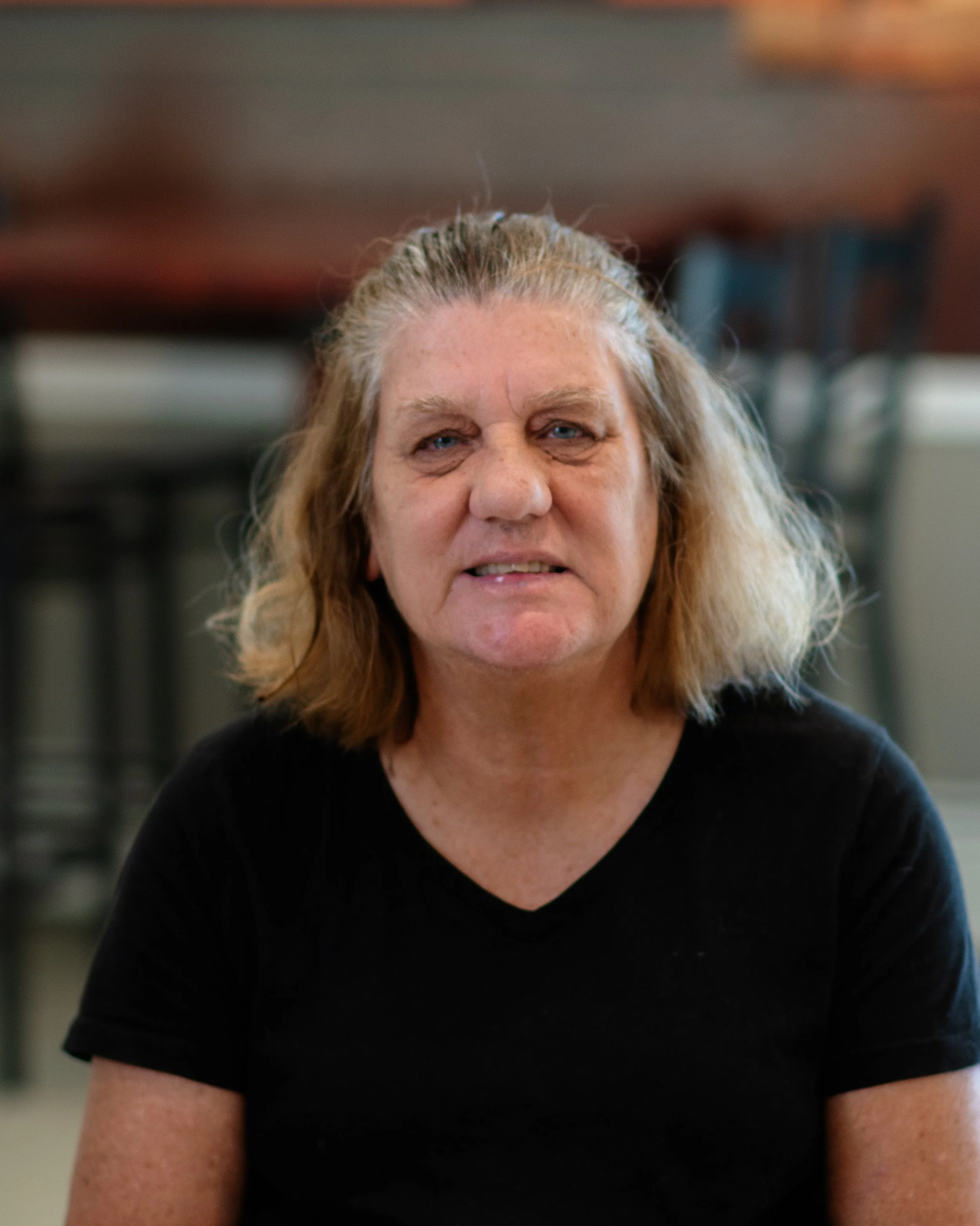 A middle-aged woman with shoulder-length blonde hair, wearing a black t-shirt, sitting indoors with a blurred background.