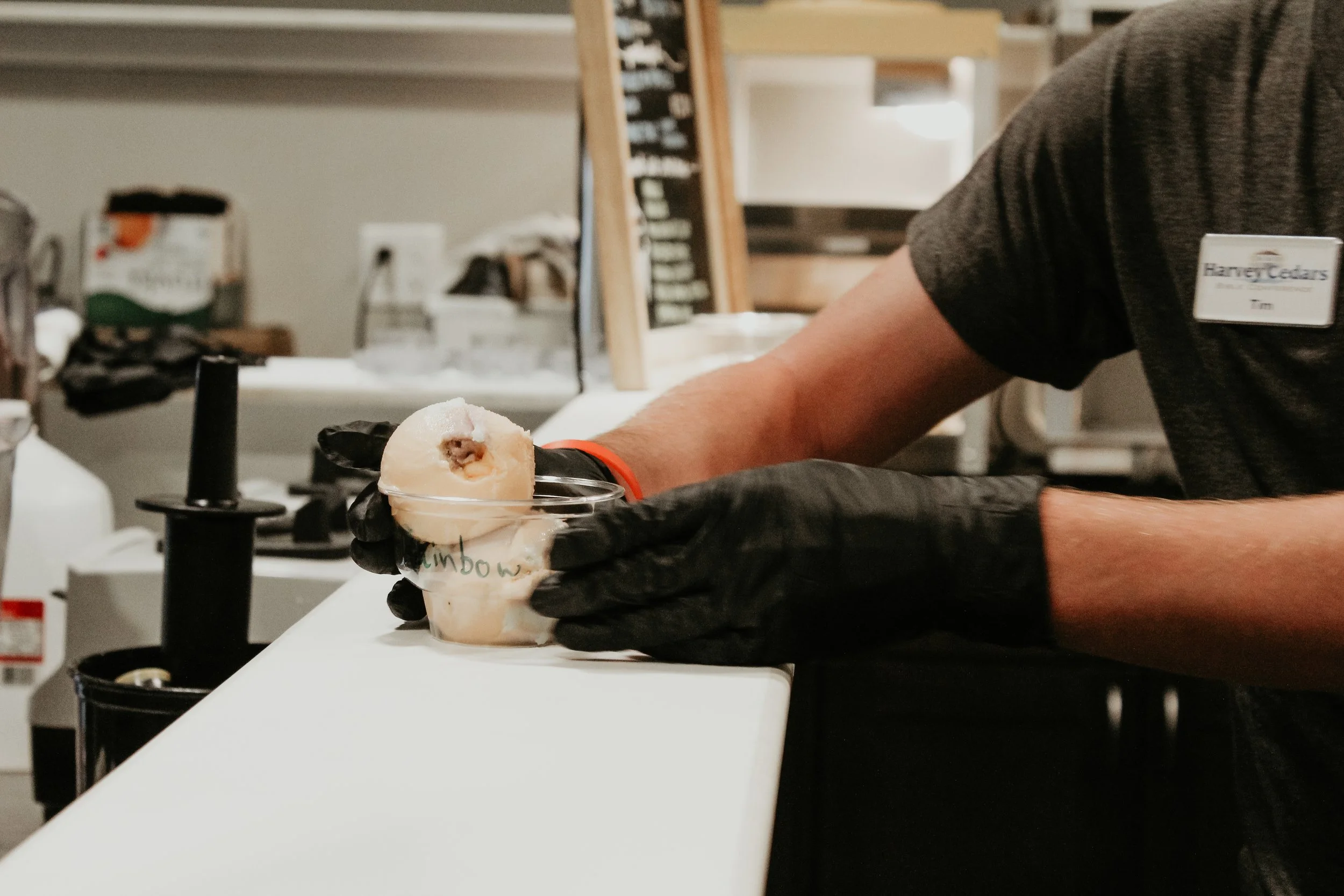 A person in black gloves placing a scoop of ice cream into a plastic cup at an ice cream shop.