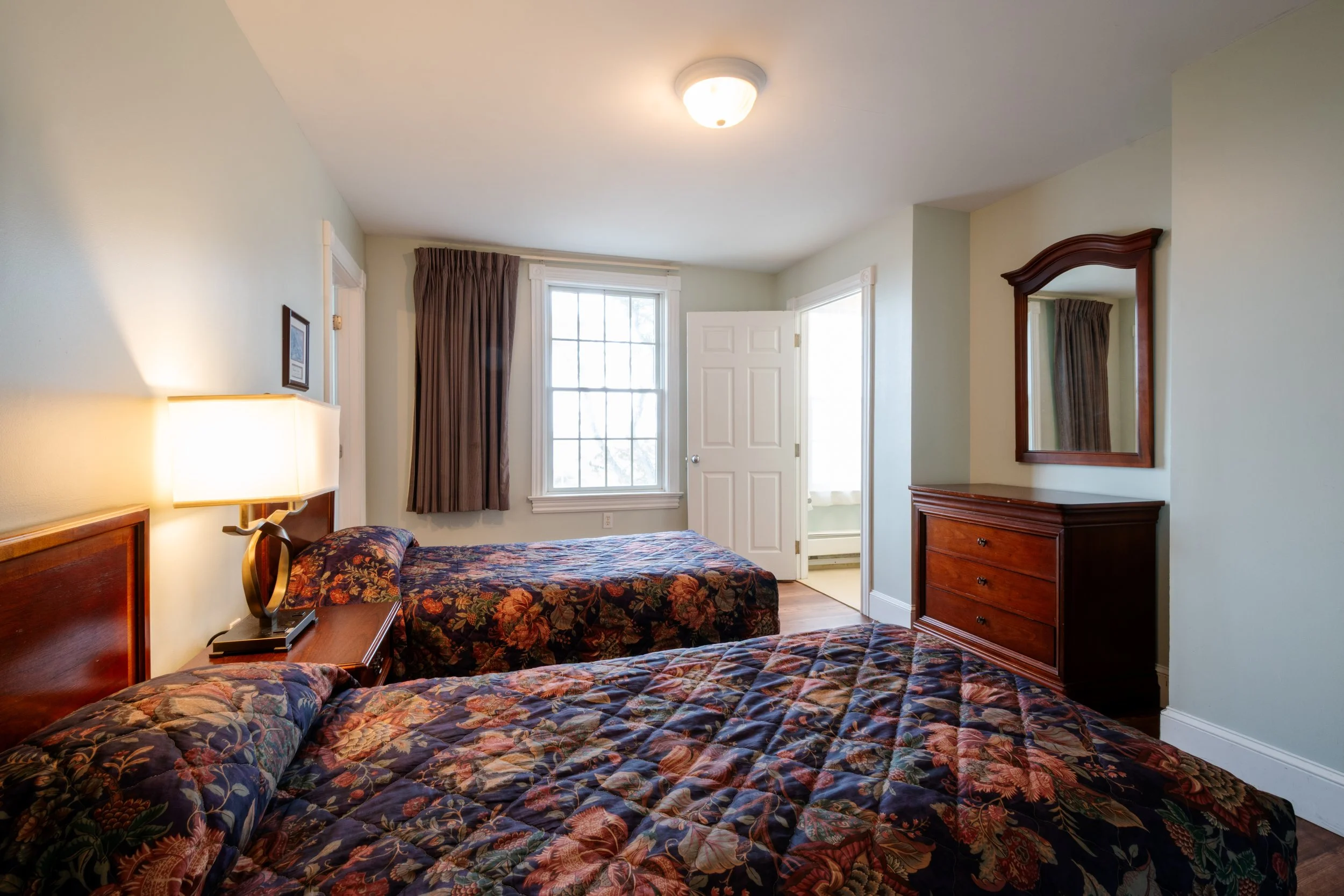 View of a bedroom with two beds with floral bedspreads, a wooden dresser with mirror, a window with curtains, and a door leading outside.