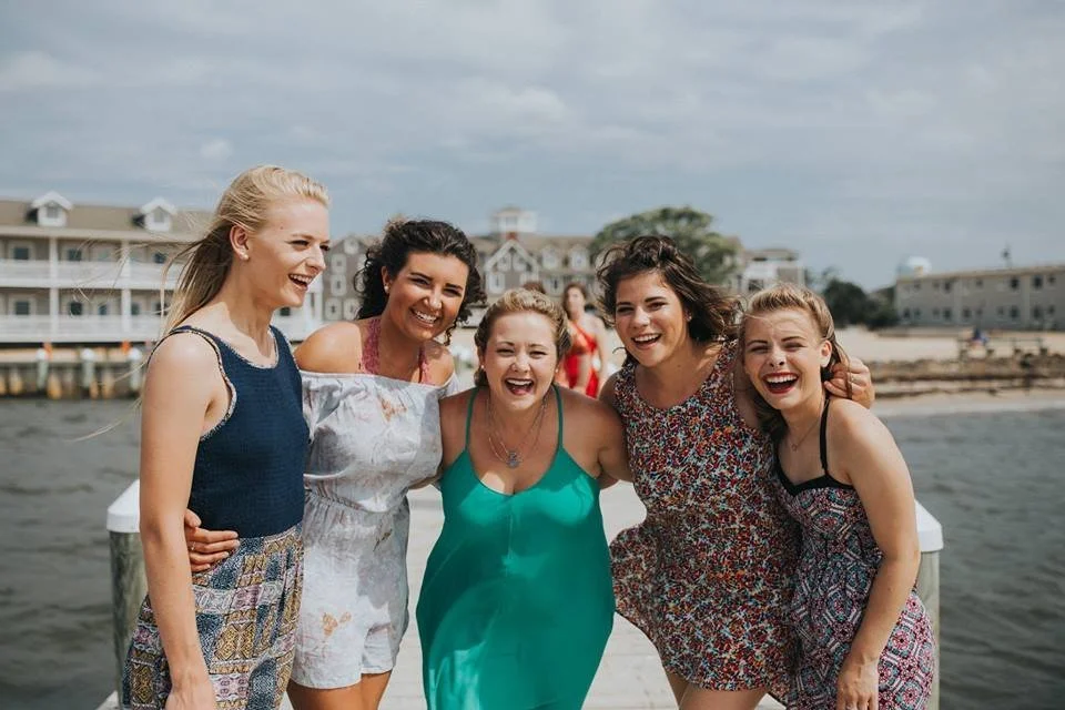 Five women standing closely together outdoors on a dock near the water, smiling and enjoying each other's company.