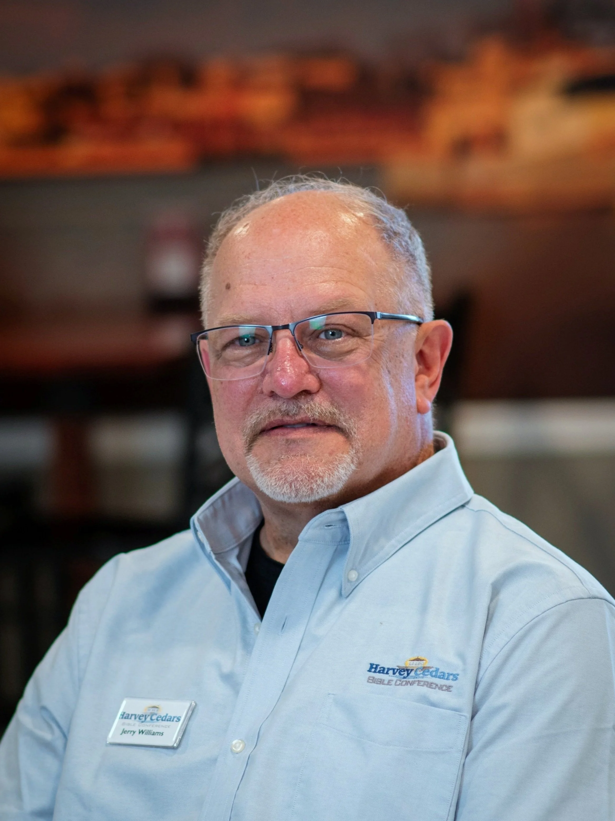 A middle-aged man with short light-colored hair, glasses, and a beard, wearing a light blue button-up shirt with a name tag, posed indoors with a blurred background.
