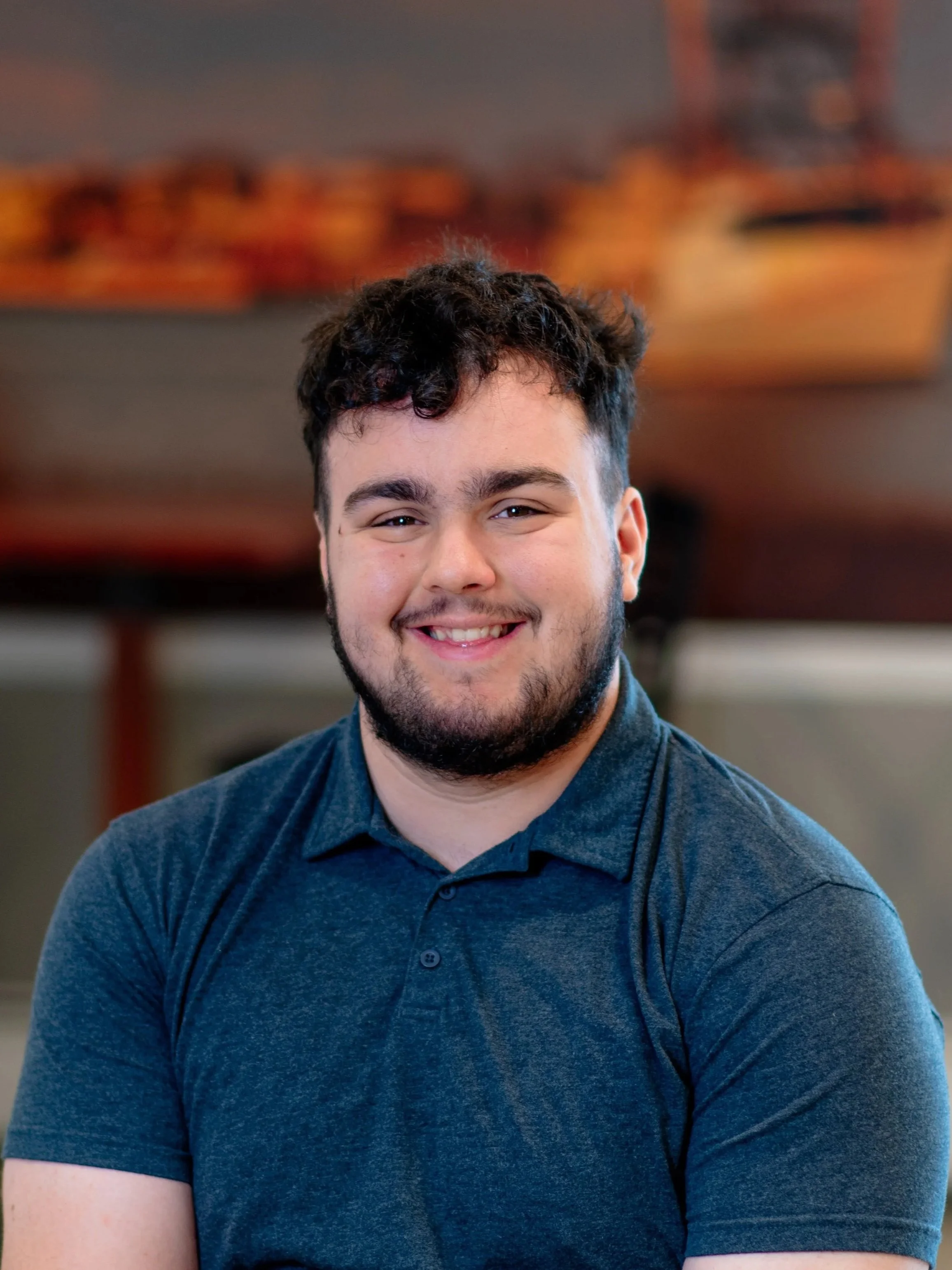 Close-up portrait of a smiling young man with dark curly hair and facial hair, wearing a dark blue polo shirt, sitting indoors with blurred background.