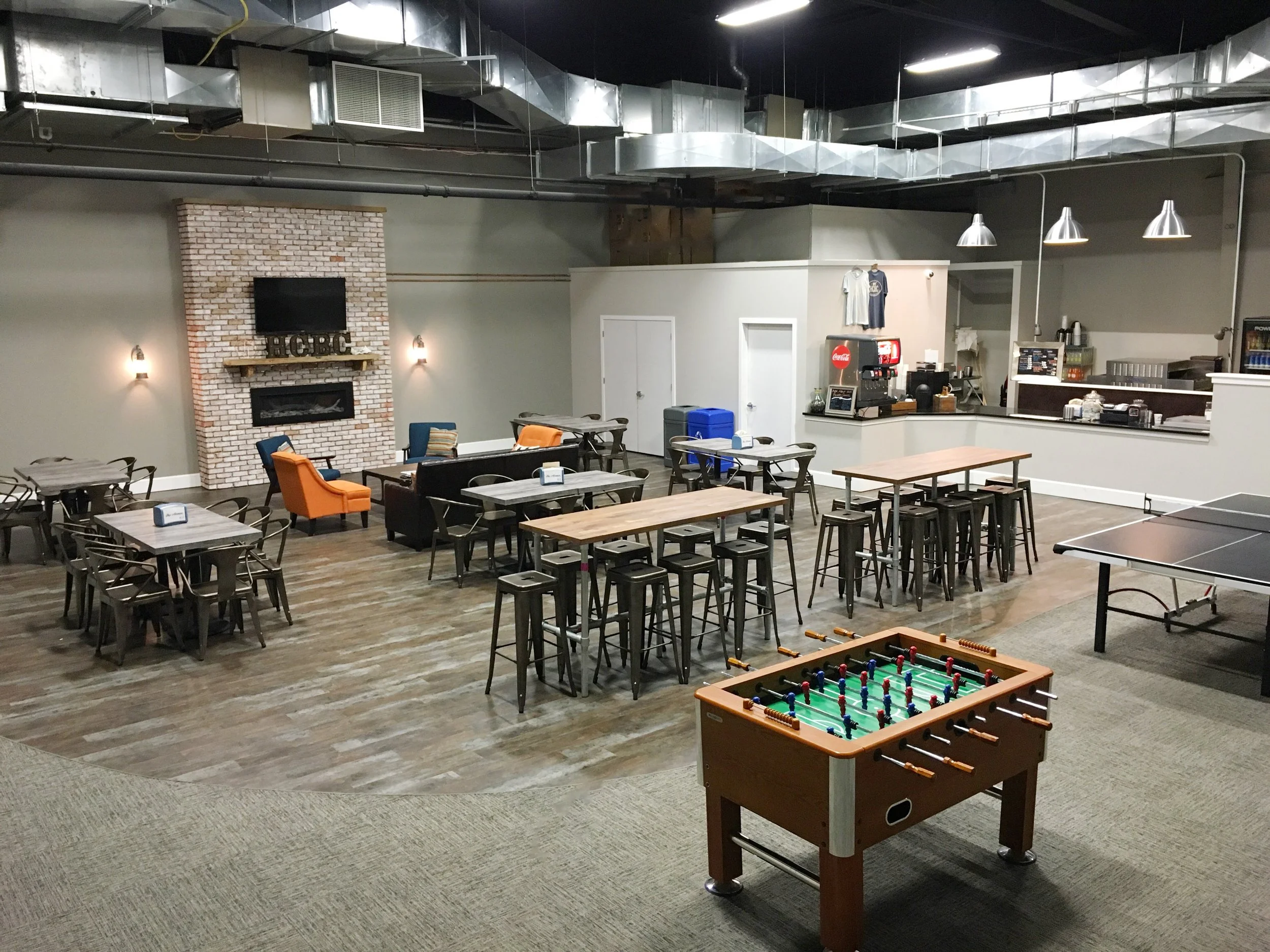 Indoor recreational space with tables, chairs, a foosball table, a ping pong table, and a lounge area featuring a brick fireplace, TV, and colorful chairs.
